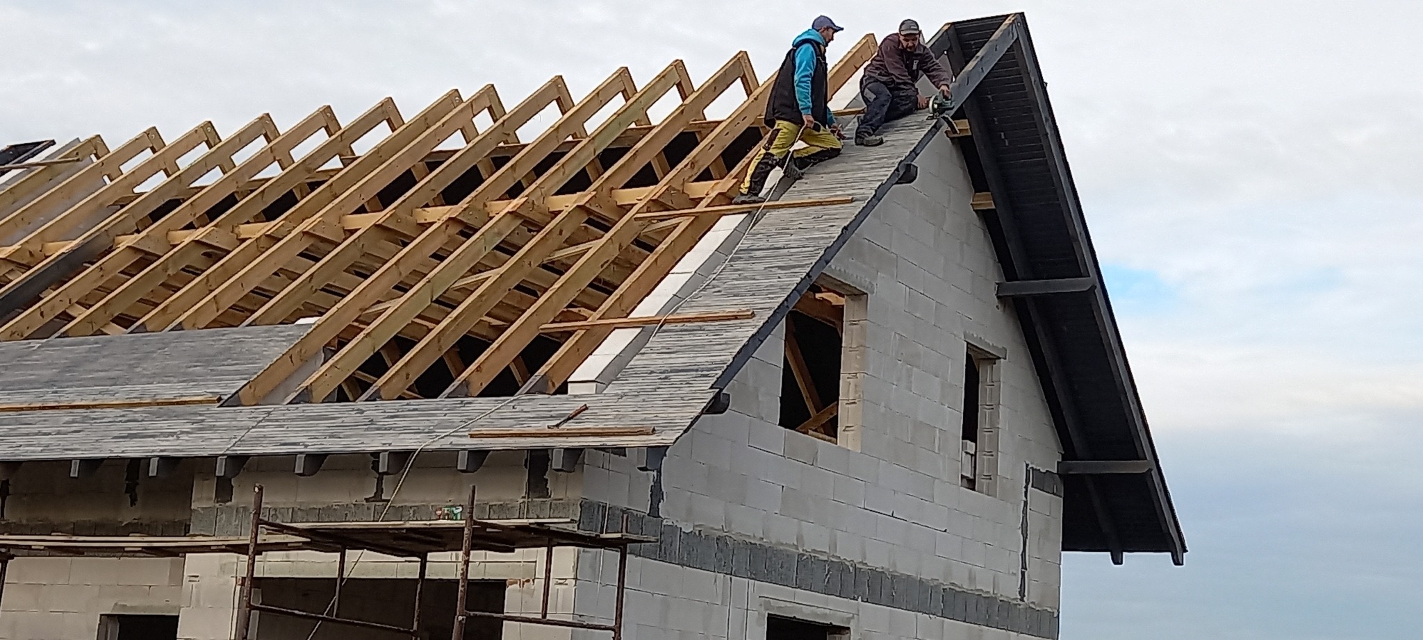 Budowa domu murowanego: dwóch robotników montuje pokrycie dachowe na konstrukcji z drewnianych krokwi, widoczny budynek z pustaków z otworami okiennymi i rusztowanie.