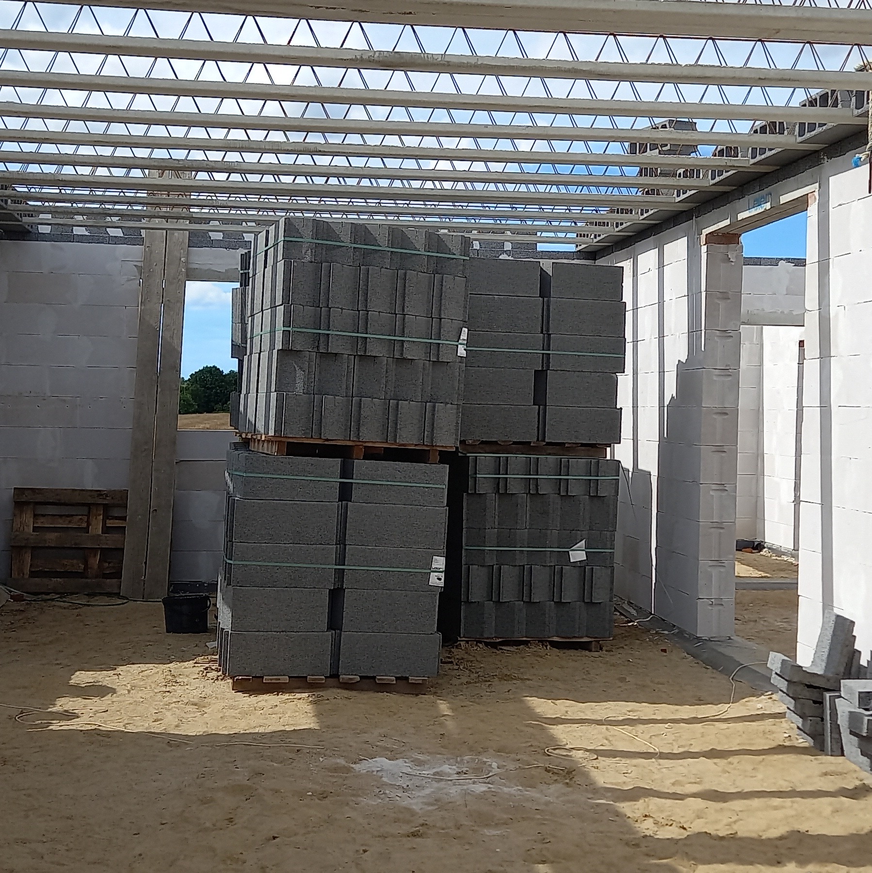 Wnętrze surowego domu w trakcie budowy, z paletami szarych bloczków ustawionymi na piaskowej podłodze, widoczne ściany z białych pustaków i stalowa konstrukcja dachu.