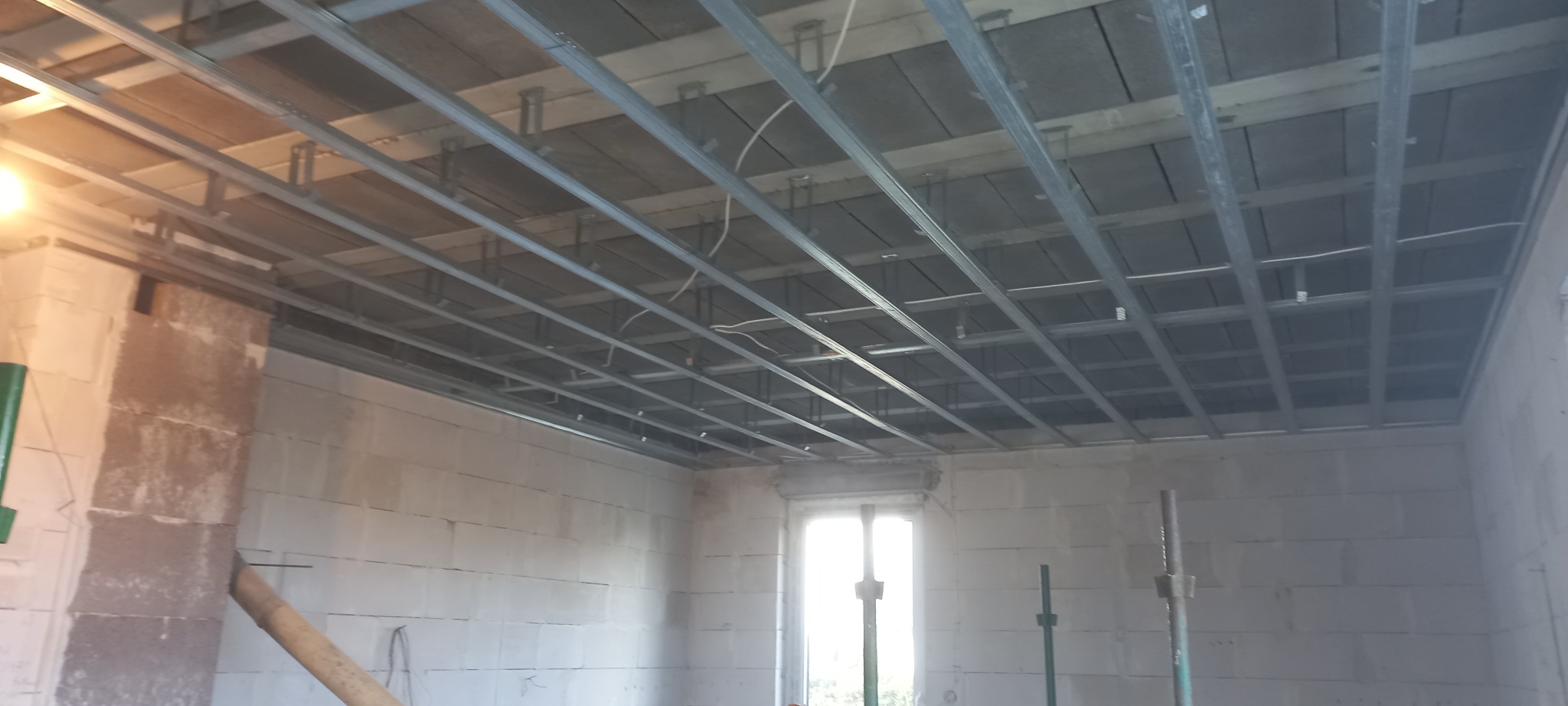 Stelaż sufitowy z profili metalowych w surowym wnętrzu z bloczków, widoczne szare płyty między profilami i podpory teleskopowe przy oknie.