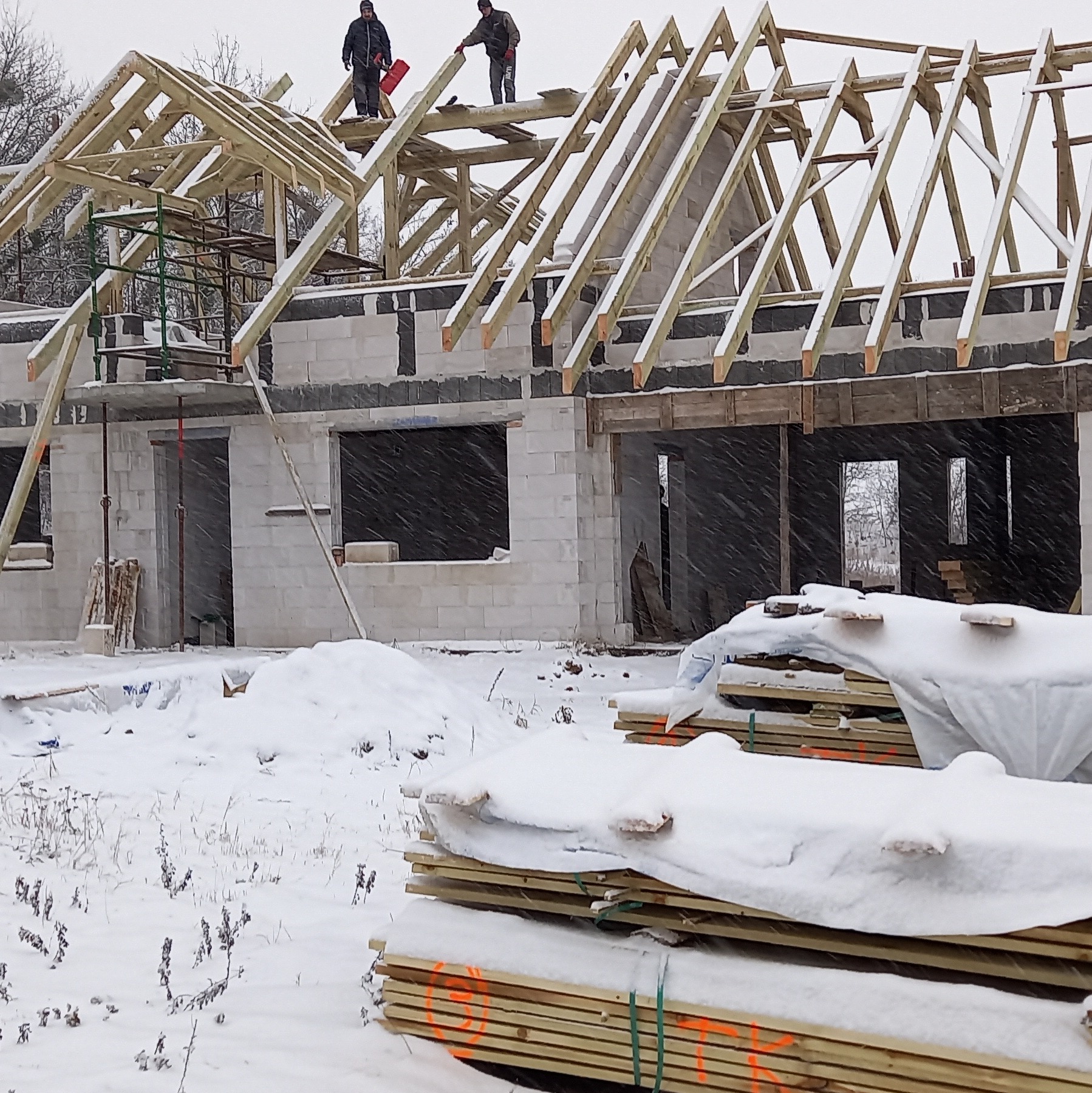 Budowa domu murowanego zimą: mur z bloczków, drewniana konstrukcja dachu w trakcie montażu, dwóch robotników na dachu, stos desek częściowo przykryty śniegiem na pierwszym planie.