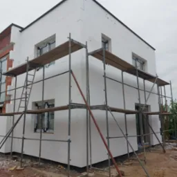 Dwukondygnacyjny budynek w trakcie ocieplania styropianem, widoczne rusztowania i fragment nieocieplonej ściany z cegły.