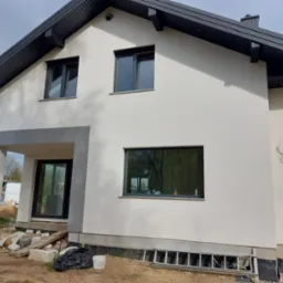 Nowoczesny dom jednorodzinny po ociepleniu i malowaniu elewacji na kolor biały, z grafitowymi oknami i dachem, widoczne przygotowania do wykończenia terenu wokół budynku.