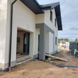 MAT-BUD Mateusz Szymański - Surowy stan elewacji domu jednorodzinnego z widocznym wejściem, fragmentem orynnowania i stertą kostki brukowej w tle. Dom w trakcie wykończenia, widoczne kable i instalacje wewnątrz.