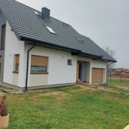 Nowoczesny dom jednorodzinny z szarą dachówką i jasną elewacją, brązowe rolety zewnętrzne, widok z zewnątrz na niezagospodarowany teren.