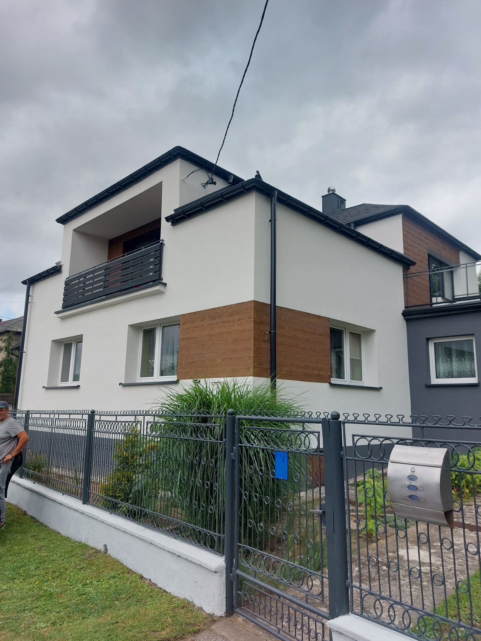 Nowoczesny dom dwukondygnacyjny z balkonami, wykończony białą i szarą elewacją z elementami drewnopodobnymi, ogrodzony metalowym płotem na niskiej podmurówce.