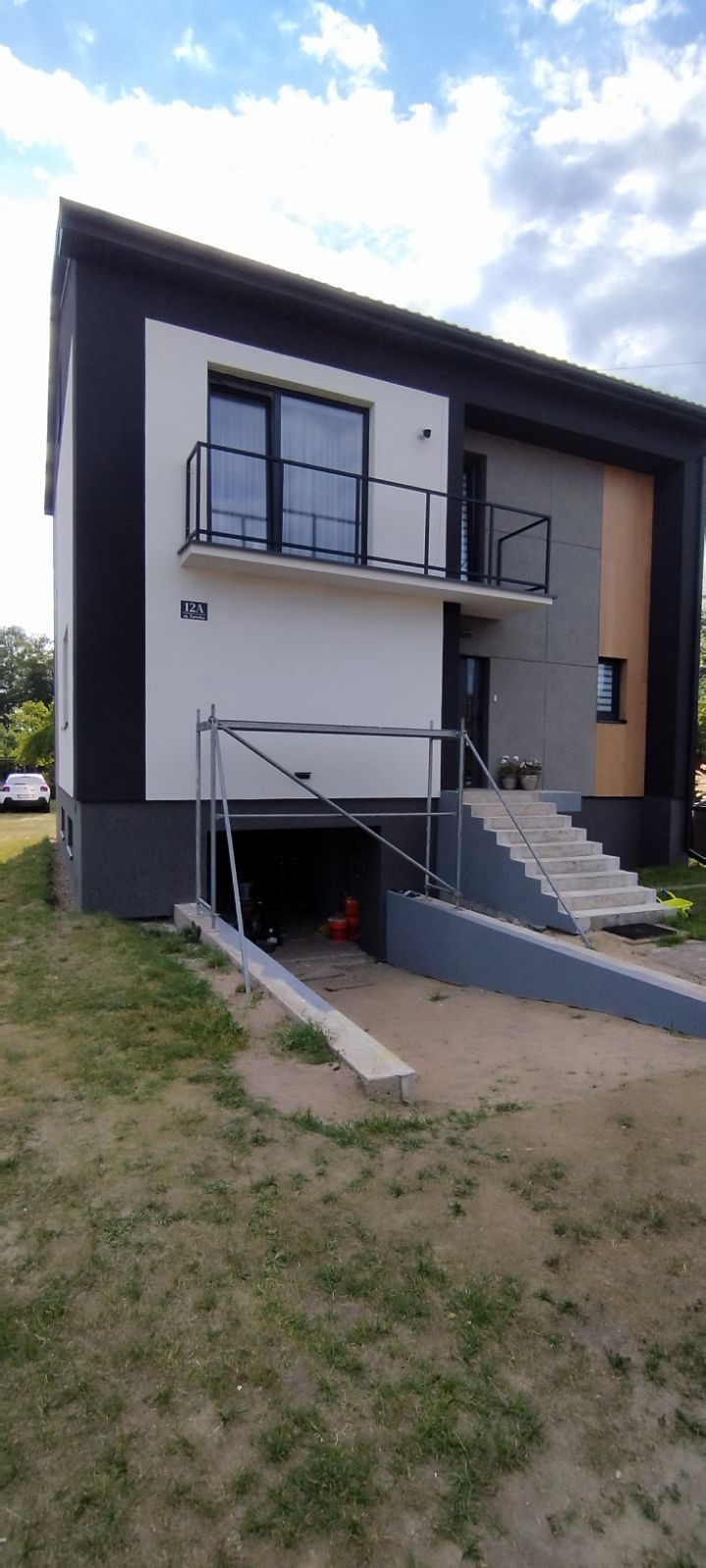 Nowoczesny dom z elewacją w odcieniach szarości, bieli i brązu, częściowo osłonięty rusztowaniem, z widocznym numerem 12A na ścianie i schodami prowadzącymi do wejścia.