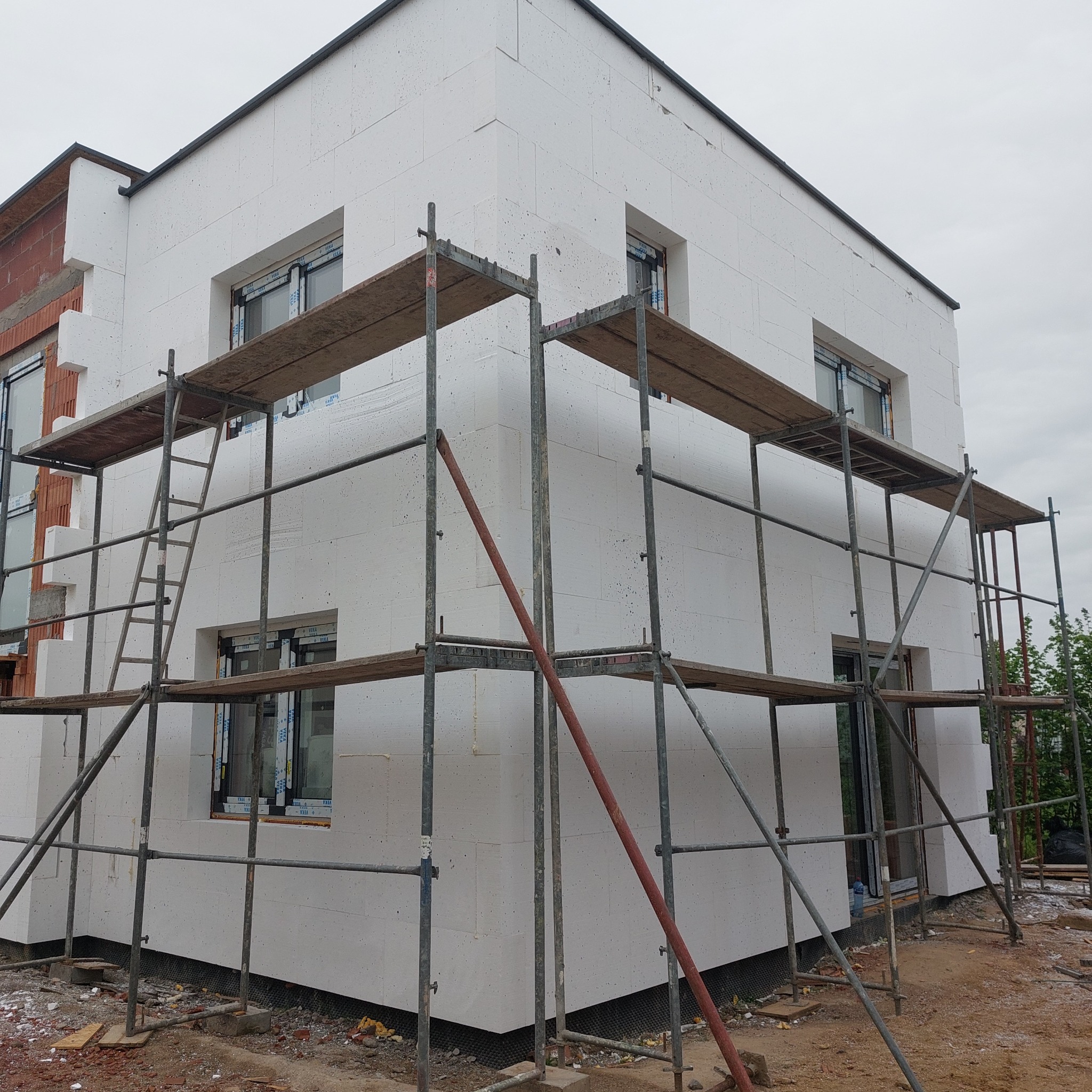 Dwukondygnacyjny budynek w trakcie ocieplania styropianem, widoczne rusztowania i fragment nieocieplonej ściany z cegły.