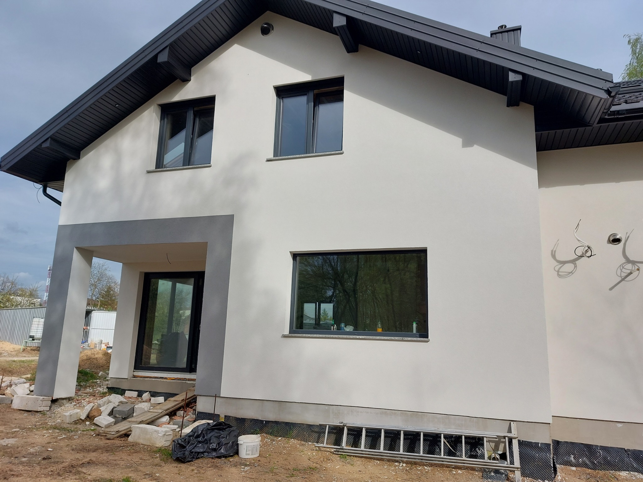 Nowoczesny dom jednorodzinny po ociepleniu i malowaniu elewacji na kolor biały, z grafitowymi oknami i dachem, widoczne przygotowania do wykończenia terenu wokół budynku.