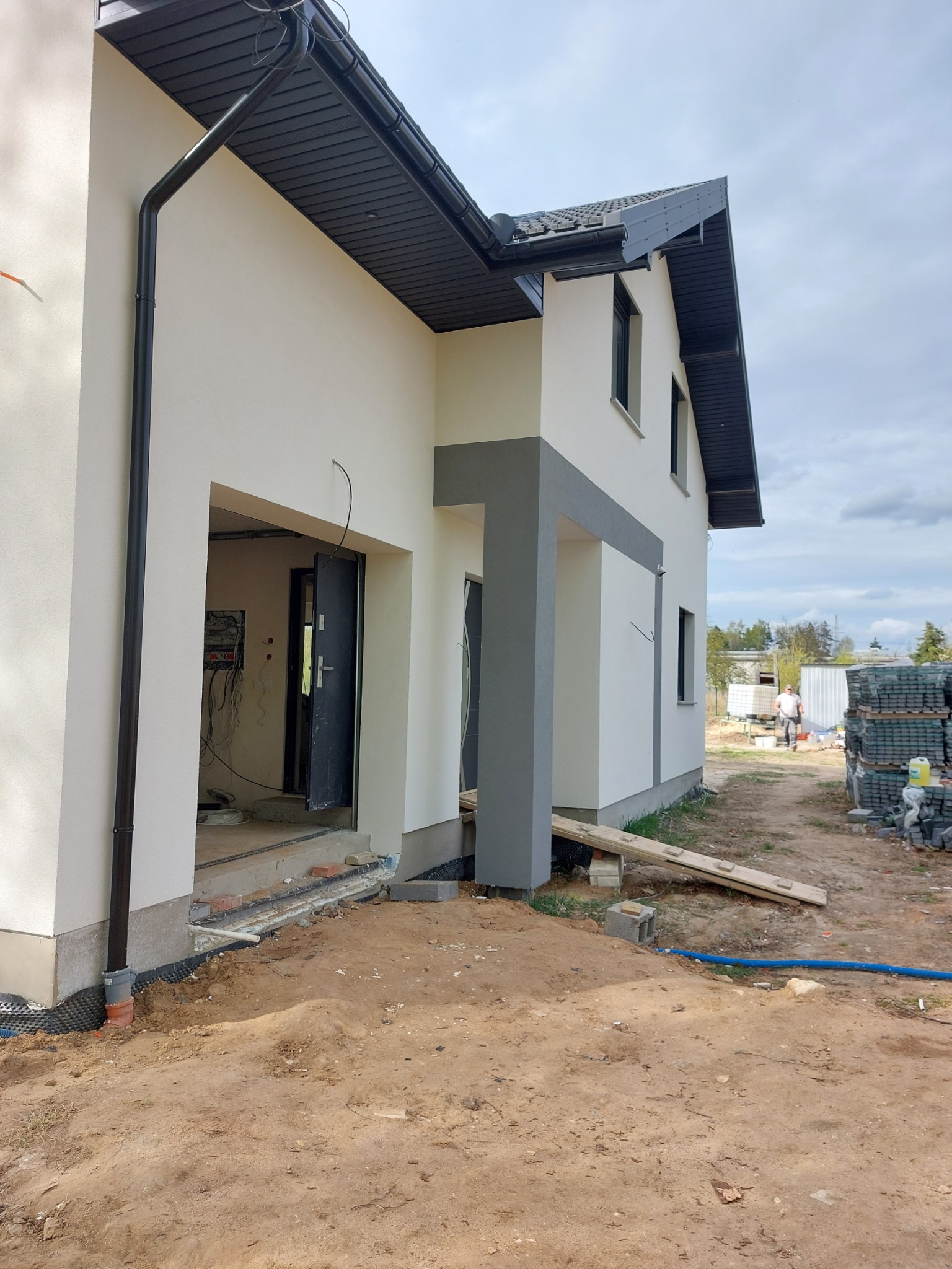 Surowy stan elewacji domu jednorodzinnego z widocznym wejściem, fragmentem orynnowania i stertą kostki brukowej w tle. Dom w trakcie wykończenia, widoczne kable i instalacje wewnątrz.