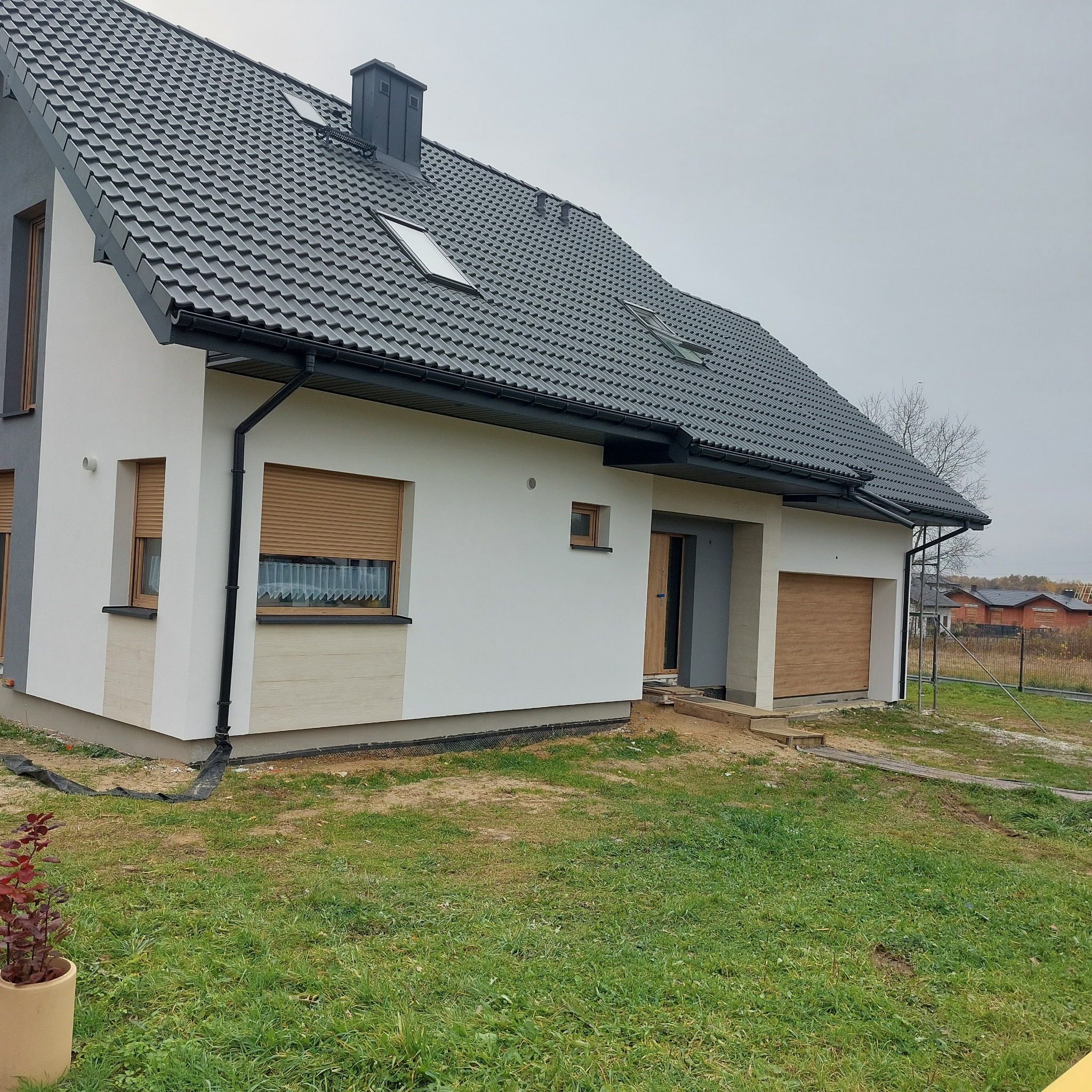 Nowoczesny dom jednorodzinny z szarą dachówką i jasną elewacją, brązowe rolety zewnętrzne, widok z zewnątrz na niezagospodarowany teren.