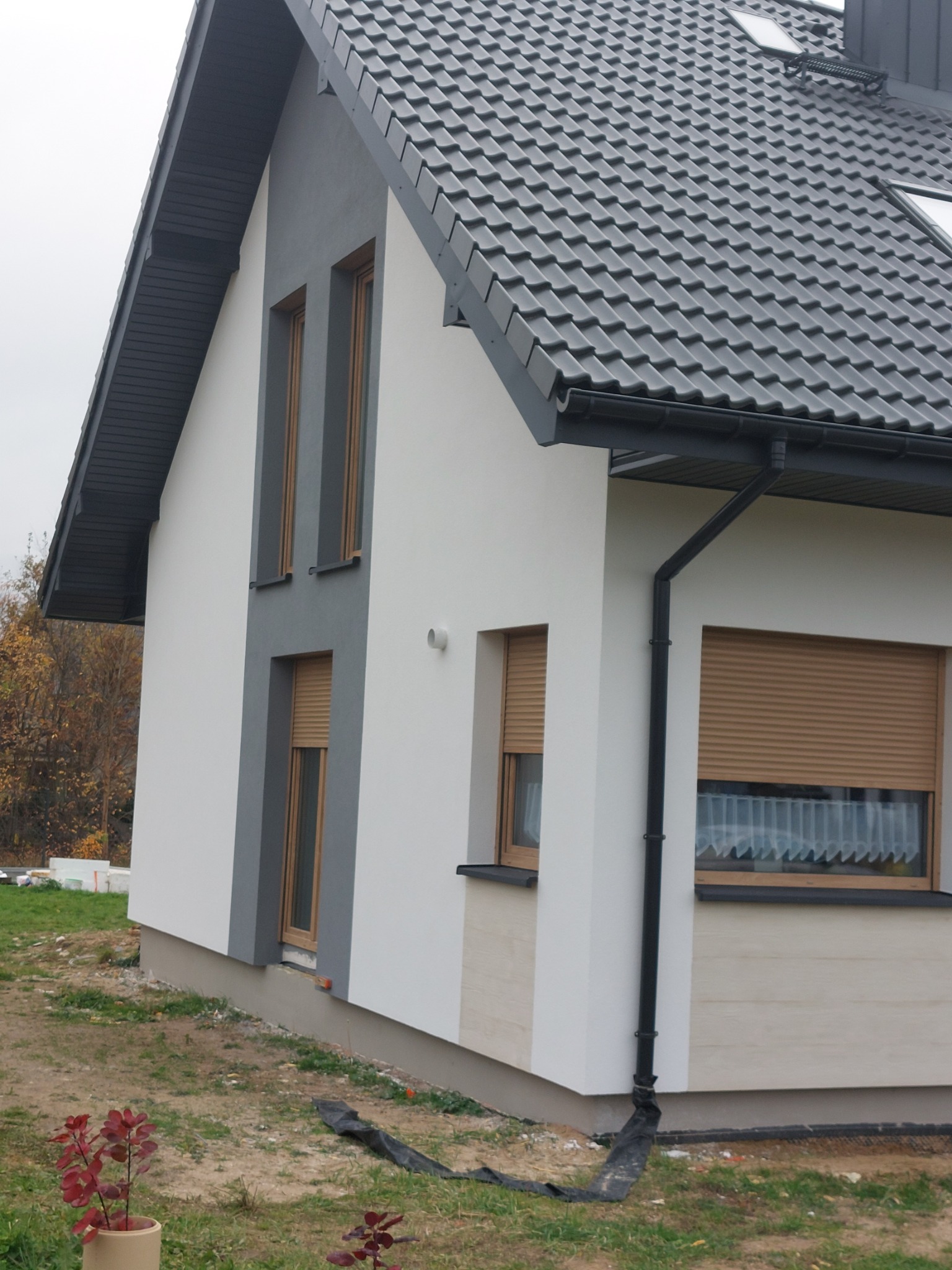 Fragment elewacji domu jednorodzinnego z szarym dachem i oknami z roletami zewnętrznymi, widoczne fragmenty ocieplenia i rynna spustowa, w tle jesienne drzewa.
