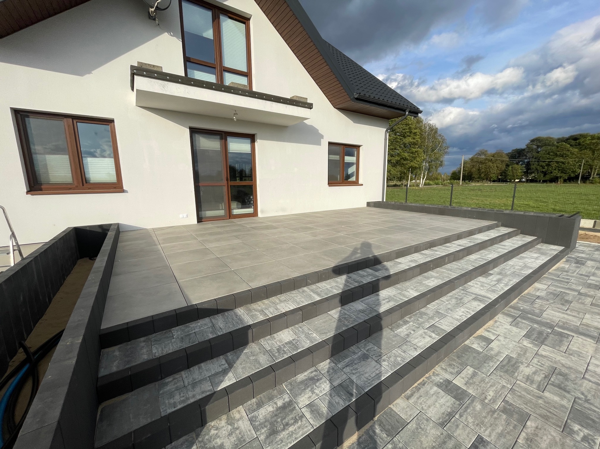 Nowoczesny taras z szarych płyt i schodami z kostki brukowej, cień fotografa na schodach, biały dom z brązowymi oknami w tle, widok na ogród i pochmurne niebo.