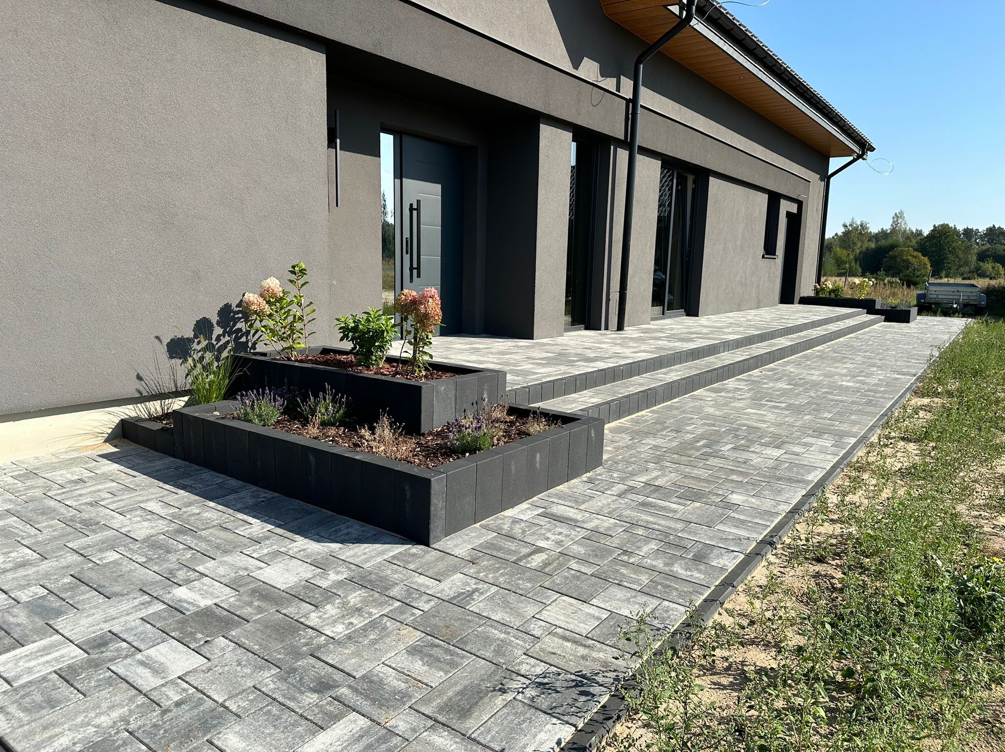 Nowoczesny dom z szarą elewacją, wejściem wyłożonym szarą kostką brukową, dwupoziomowymi, czarnymi donicami z roślinami i kwiatami, oraz widocznym fragmentem ogrodu.