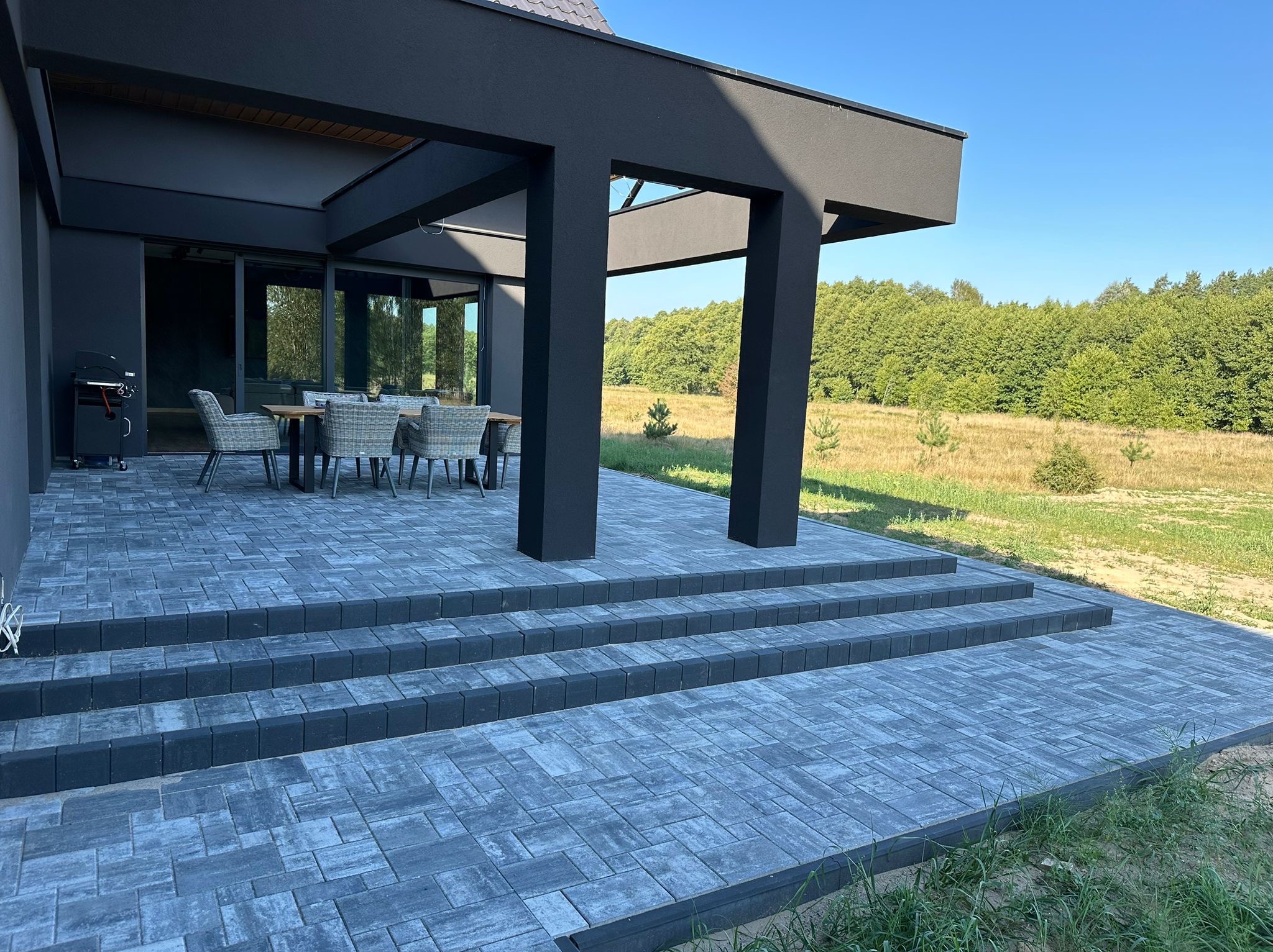 Nowoczesny taras z szarej kostki brukowej prowadzący do domu z czarną elewacją, widok na zielony ogród i las w tle.