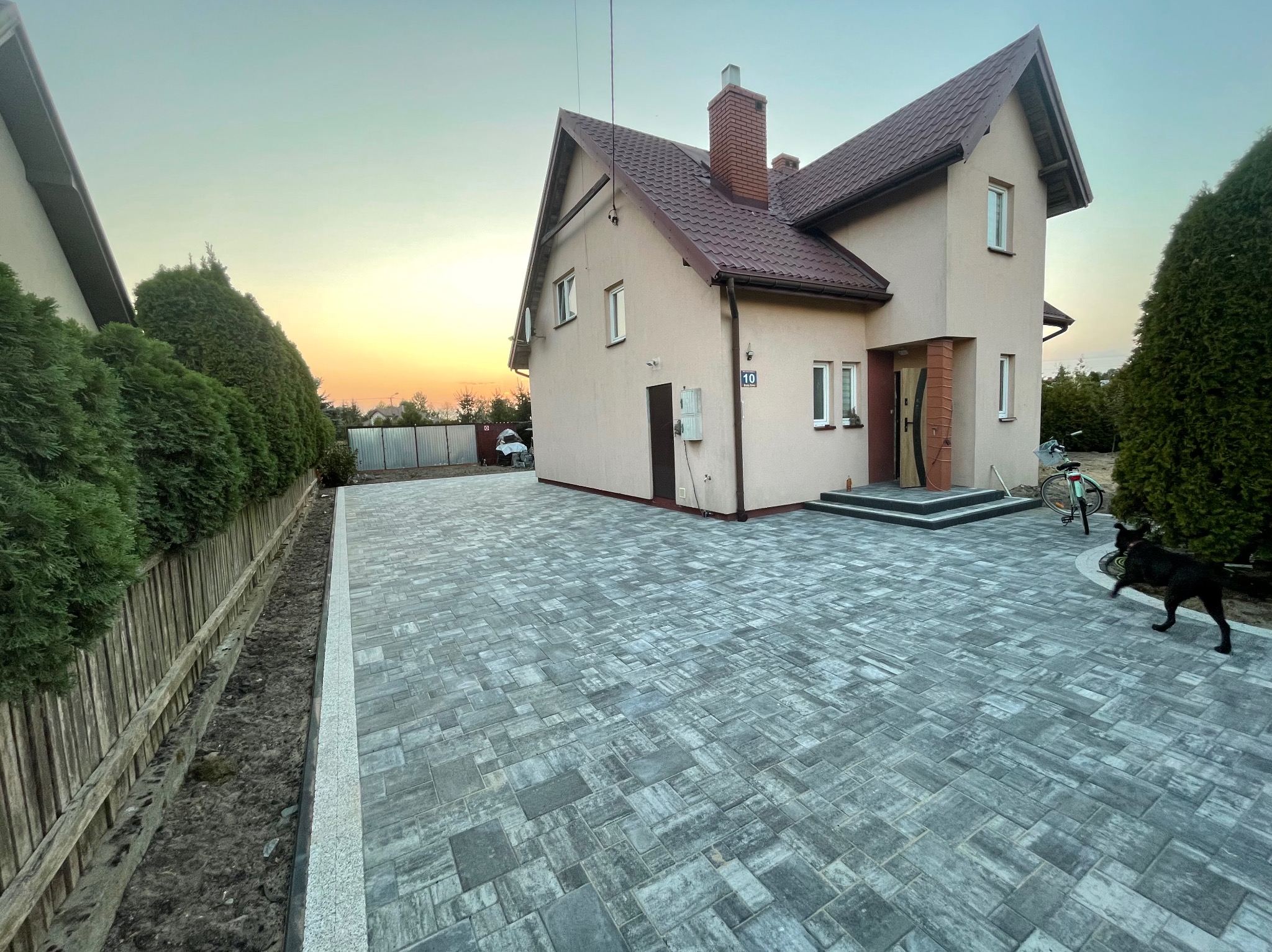 Nowo położona, szara kostka brukowa przed domem z brązowym dachem, widoczny fragment ogrodu z tujami i czarny pies bawiący się piłką.