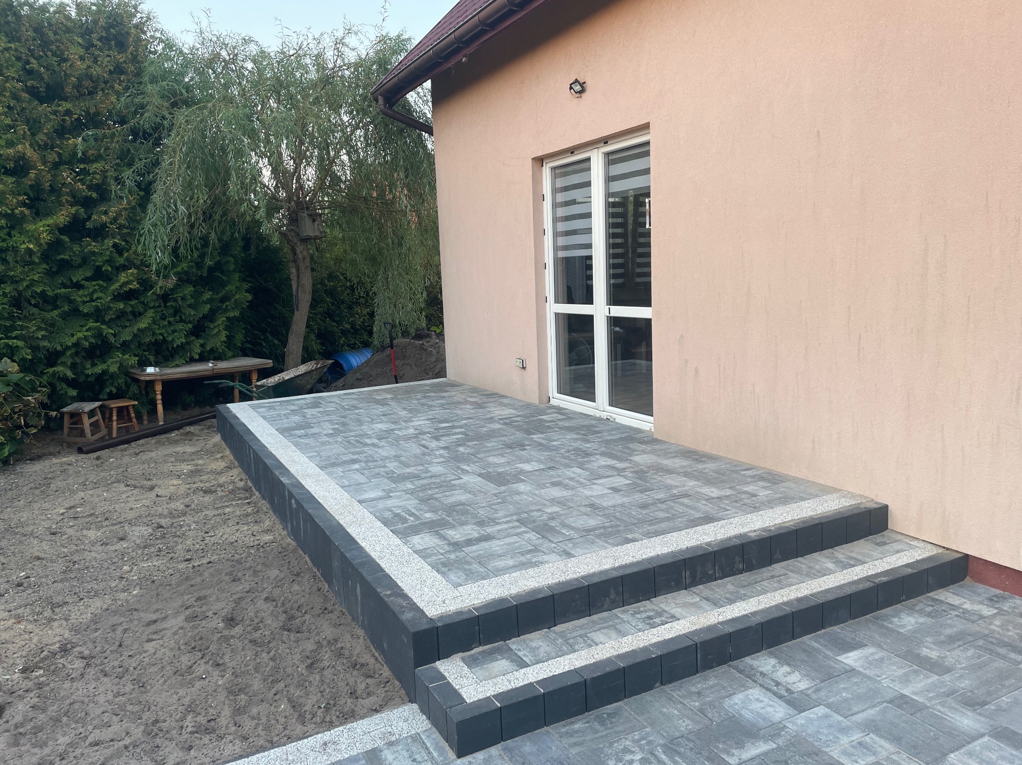 Nowoczesny taras z szarej kostki brukowej z dwoma stopniami, obrzeża z drobnego grysu, przylegający do budynku z beżową elewacją i białymi drzwiami balkonowymi, w tle drzewo.
