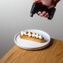 Prostokątna tarta z kremem przypalanym palnikiem cukierniczym na białym talerzu, leżącym na drewnianym stole.