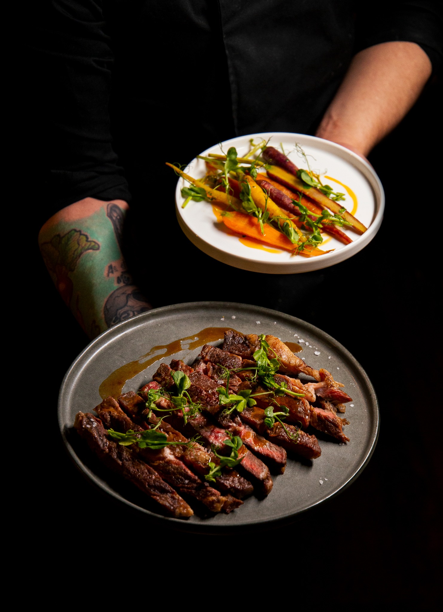 Elegancko podane danie: pokrojony stek z sosem i mikro-ziołami na szarym talerzu oraz kolorowe marchewki na białym talerzu, trzymane przez osobę z tatuażem na ramieniu, na czarnym tle.
