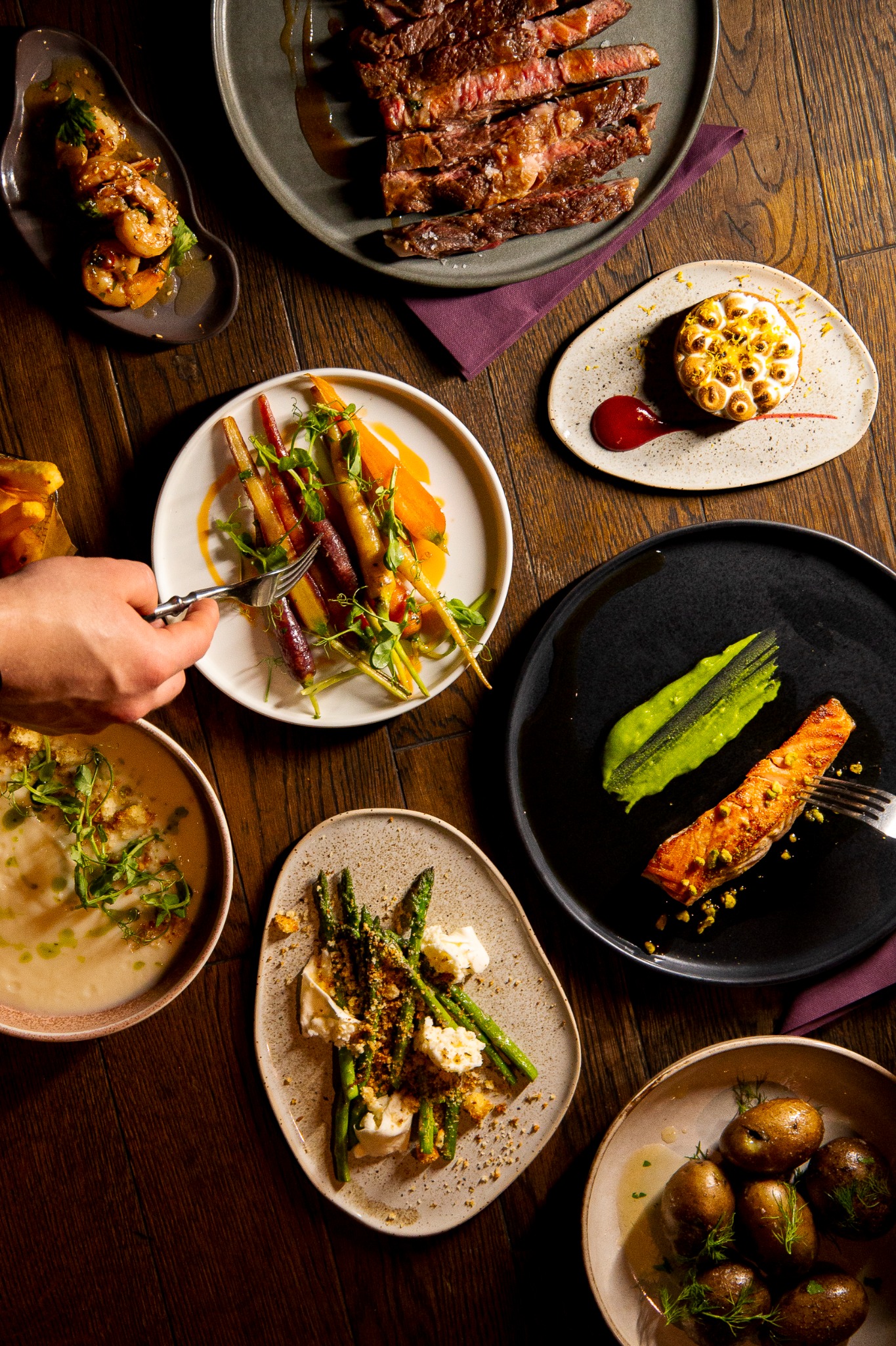 Widok z góry na stół z różnorodnymi daniami: stek, krewetki, kolorowe marchewki, zupa krem, szparagi z serem burrata, pieczony łosoś z zielonym puree i pieczone ziemniaki.