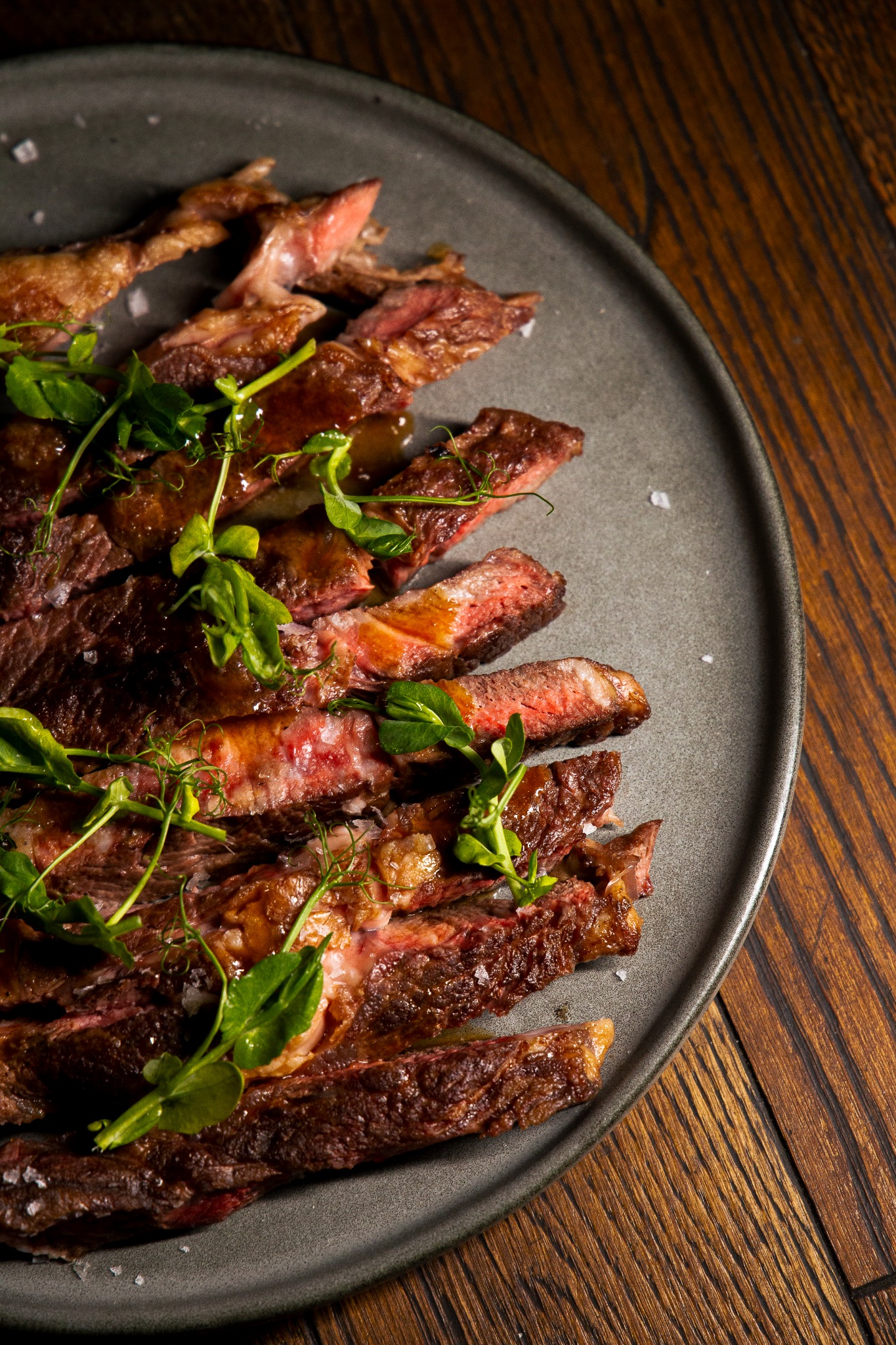 Plasterki średnio wysmażonej wołowiny, polane sosem i udekorowane świeżymi pędami groszku, ułożone na szarym talerzu z solą morską, na tle drewnianego blatu.