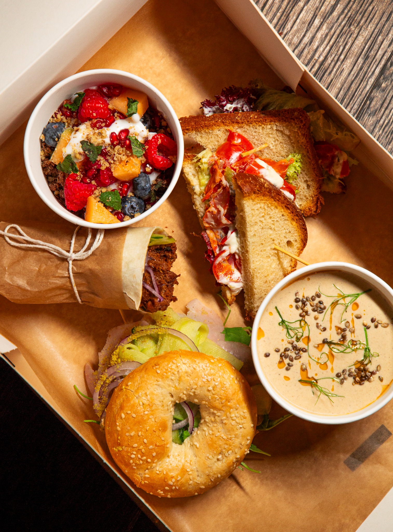 Zestaw lunchowy w pudełku: sałatka owocowa z granatem, kanapka na szpilce, wrap w papierze, bajgiel z warzywami i zupa krem z pestkami.