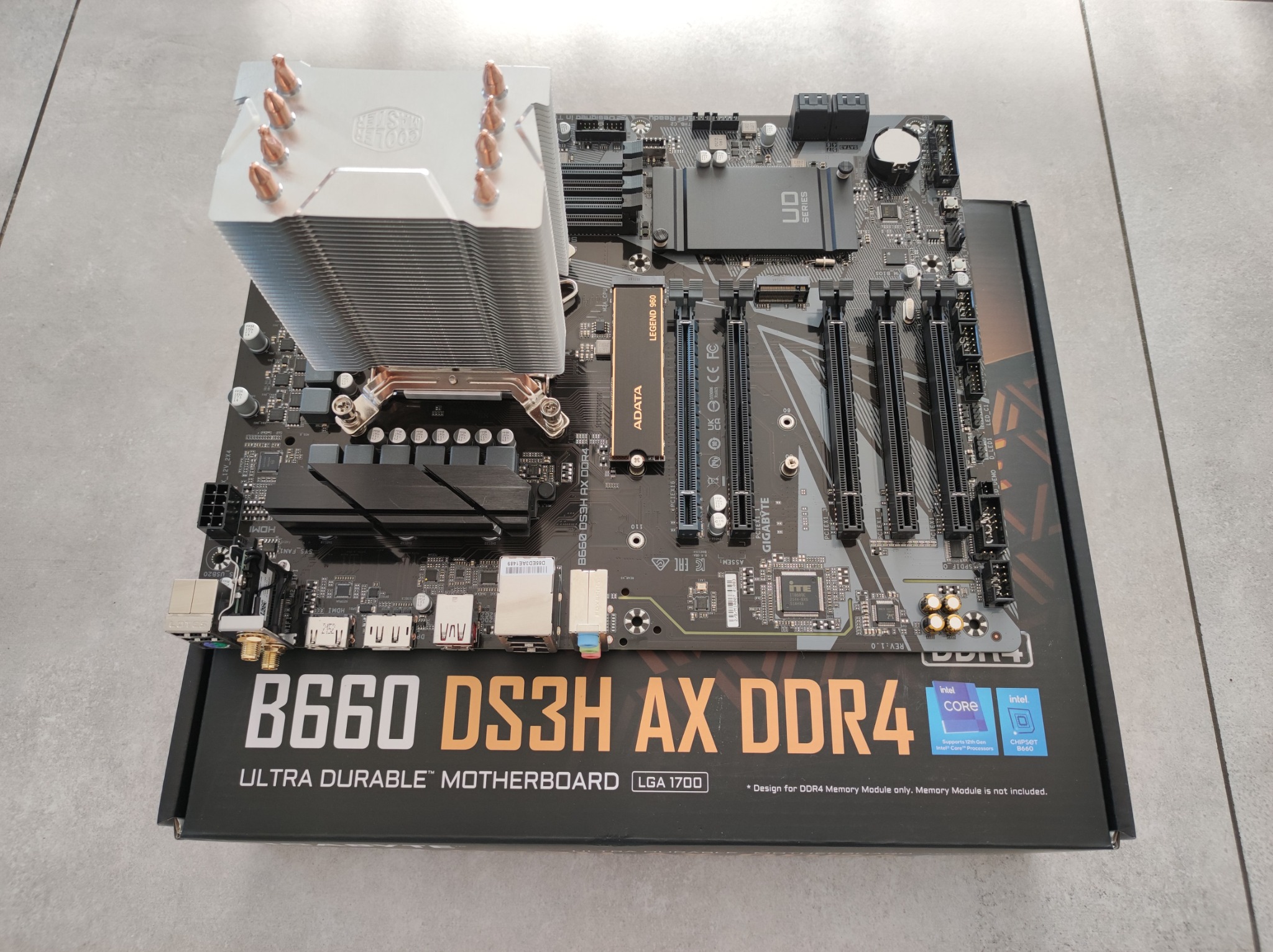 Płyta główna Gigabyte B660 DS3H AX DDR4 z zamontowanym chłodzeniem CPU Cooler Master, leżąca na pudełku na tle płytek podłogowych.