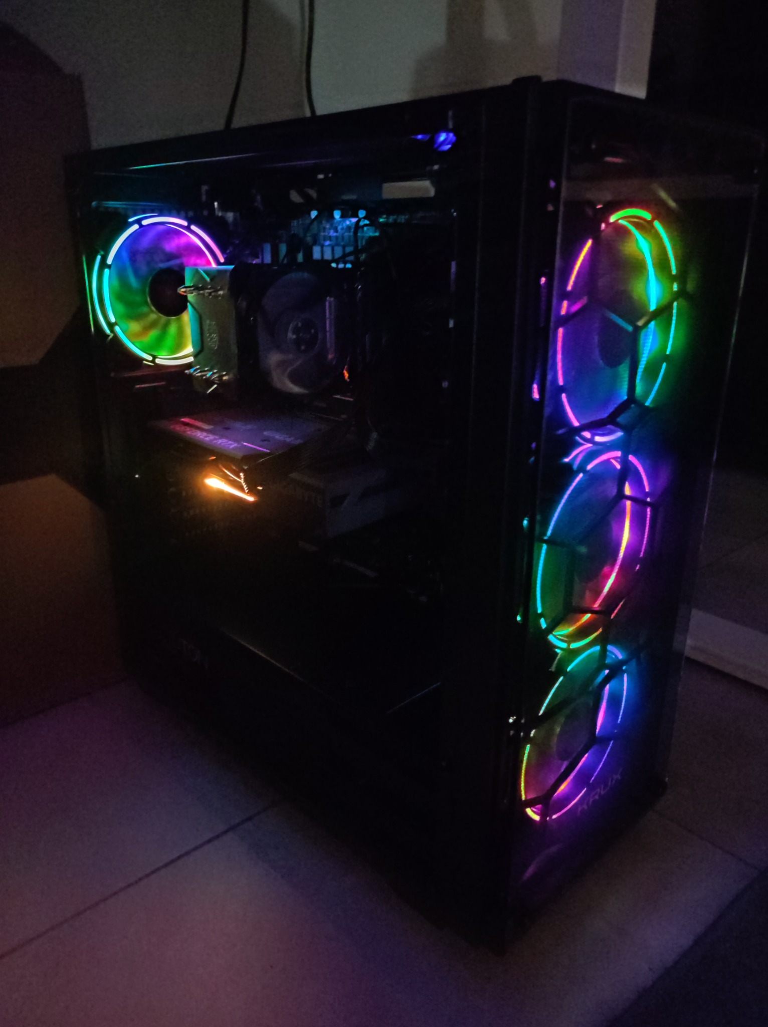 Komputer stacjonarny z przezroczystą obudową, podświetlany wentylatorami RGB i elementami wewnętrznymi, w słabym oświetleniu, widoczny napis KRUX na obudowie.