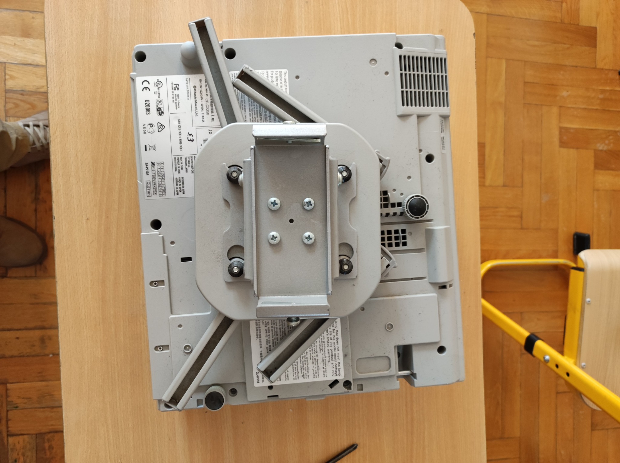 Szary projektor Hitachi zamontowany na metalowym uchwycie sufitowym, leżący na drewnianym stole. Widoczne śruby mocujące i fragment żółtego krzesła w tle.