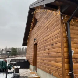 Drewniana elewacja domu z oświetleniem dekoracyjnym, widoczne elementy wykończeniowe dachu i otoczenie w zimowej scenerii.