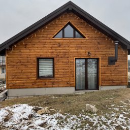 Szymon Wątor SW-BUD Usługi Wykończeniowo-Budowlane - Drewniany dom z brązowym dachem i dekoracyjnymi lampkami, widoczny komin i okno z żaluzjami, częściowo ośnieżony teren.