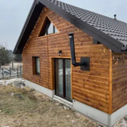 Drewniany dom z brązową blachodachówką, oświetleniem dekoracyjnym i czarnym kominem zewnętrznym, widok z zewnątrz na tle pochmurnego nieba.