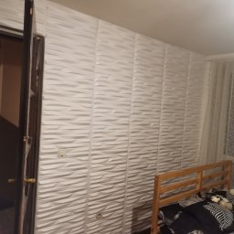 Szymon Wątor SW-BUD Usługi Wykończeniowo-Budowlane - Ściana wykończona białymi panelami 3D z włącznikiem i gniazdkiem elektrycznym, widoczna drewniana rama łóżka i fragment otwartych drzwi.