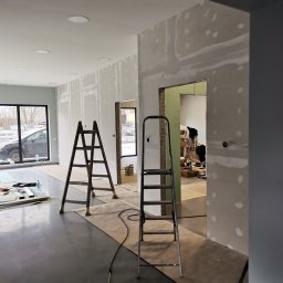 Szymon Wątor SW-BUD Usługi Wykończeniowo-Budowlane - Wnętrze w trakcie wykańczania: ściany pokryte płytami gipsowo-kartonowymi, widoczne łączenia szpachlowane, dwie drabiny, narzędzia i materiały budowlane na podłodze, duże okno z widokiem na zewnątrz.