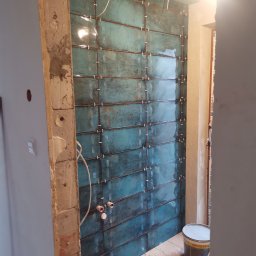 Szymon Wątor SW-BUD Usługi Wykończeniowo-Budowlane - Ściana w trakcie układania turkusowych płytek, widoczne krzyżyki dystansowe, wystające rury instalacji hydraulicznej, surowe wykończenie ościeżnicy i wiadro z zaprawą.