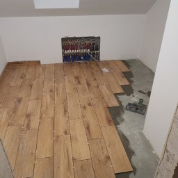 Szymon Wątor SW-BUD Usługi Wykończeniowo-Budowlane - Układanie drewnopodobnych płytek podłogowych z użyciem krzyżyków dystansowych, widoczna instalacja grzewcza w ścianie i narzędzia glazurnicze.