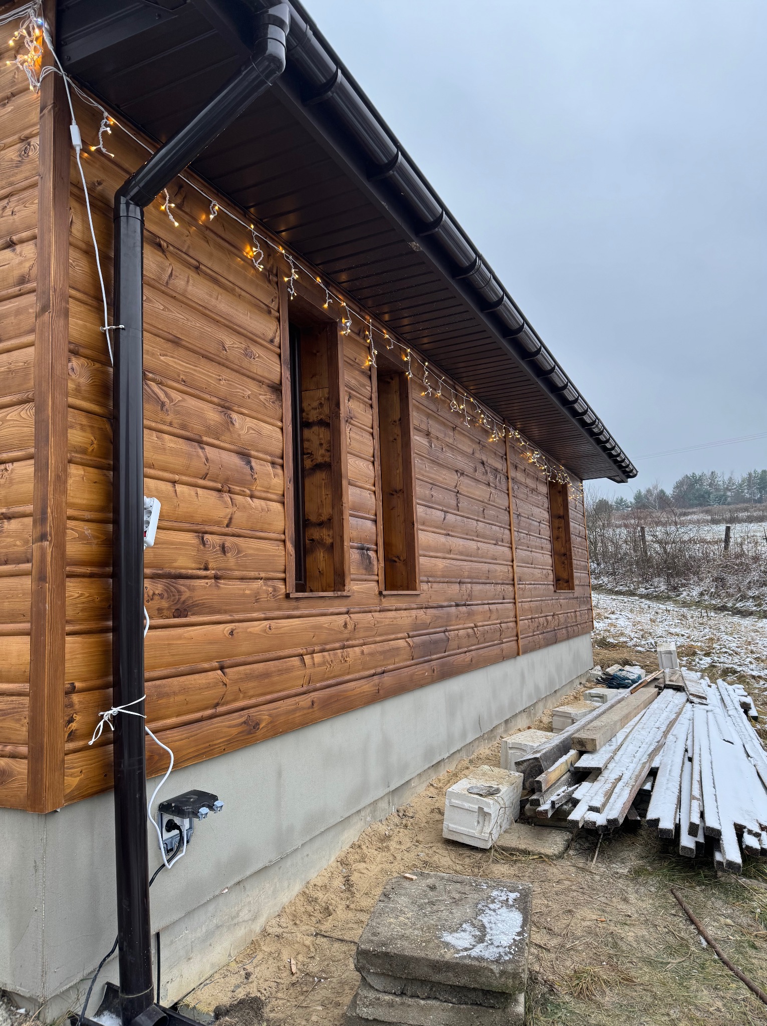 Drewniana elewacja domu z widocznymi niezabudowanymi otworami okiennymi, oświetlona girlandą świetlną, z brązowymi rynnami i widocznym przyłączem elektrycznym na zewnątrz, oraz składowanymi...