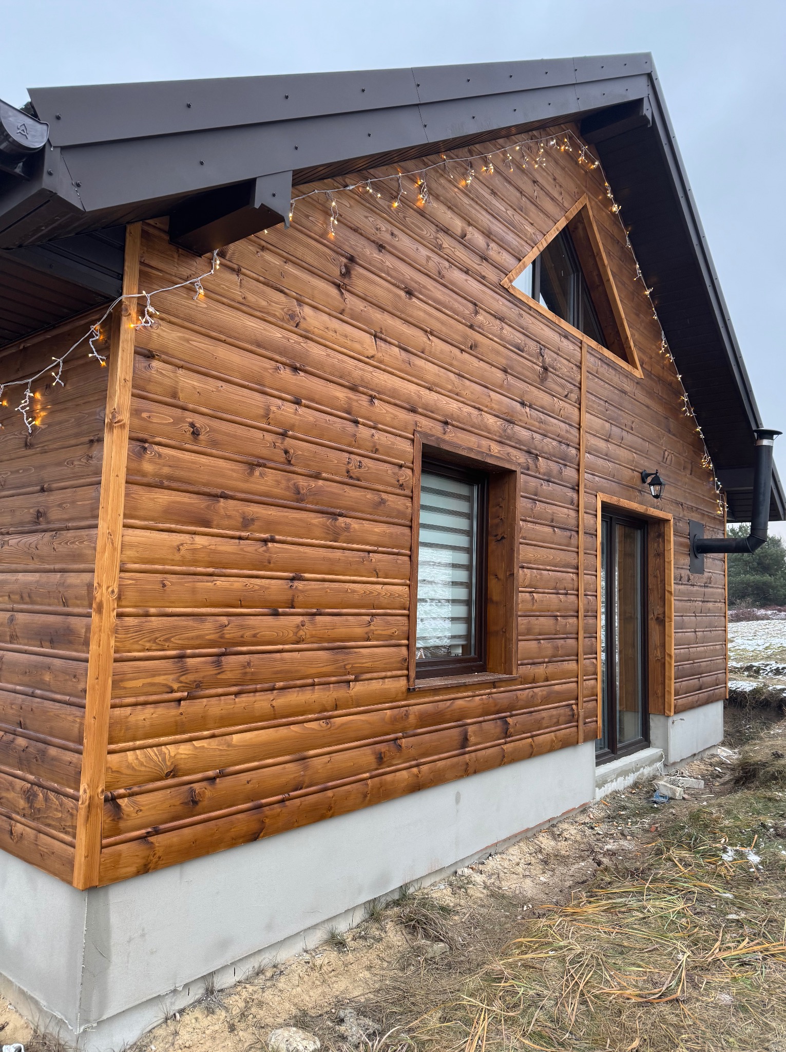 Nowoczesny dom z drewnianą elewacją w odcieniach brązu, ozdobiony lampkami, z widocznym kominem i oknem z roletami, na tle pochmurnego nieba.