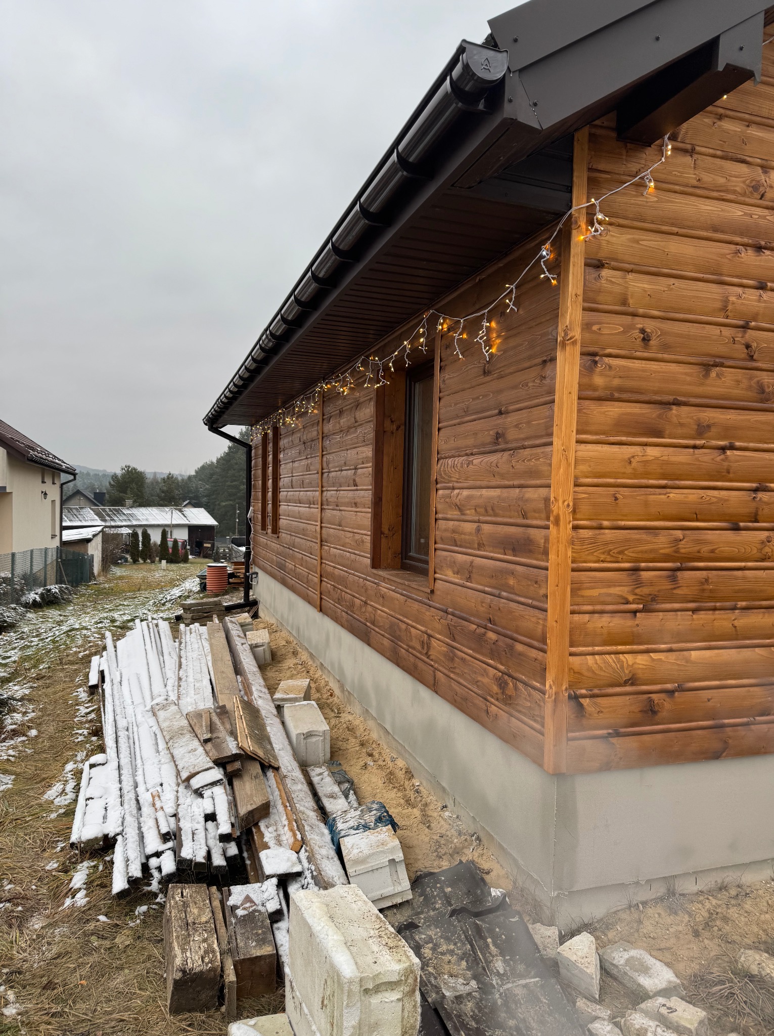 Ściana zewnętrzna drewnianego domu z oknami, udekorowana lampkami, z widocznym fundamentem i stertą desek oraz bloczków na ziemi w pochmurny dzień.