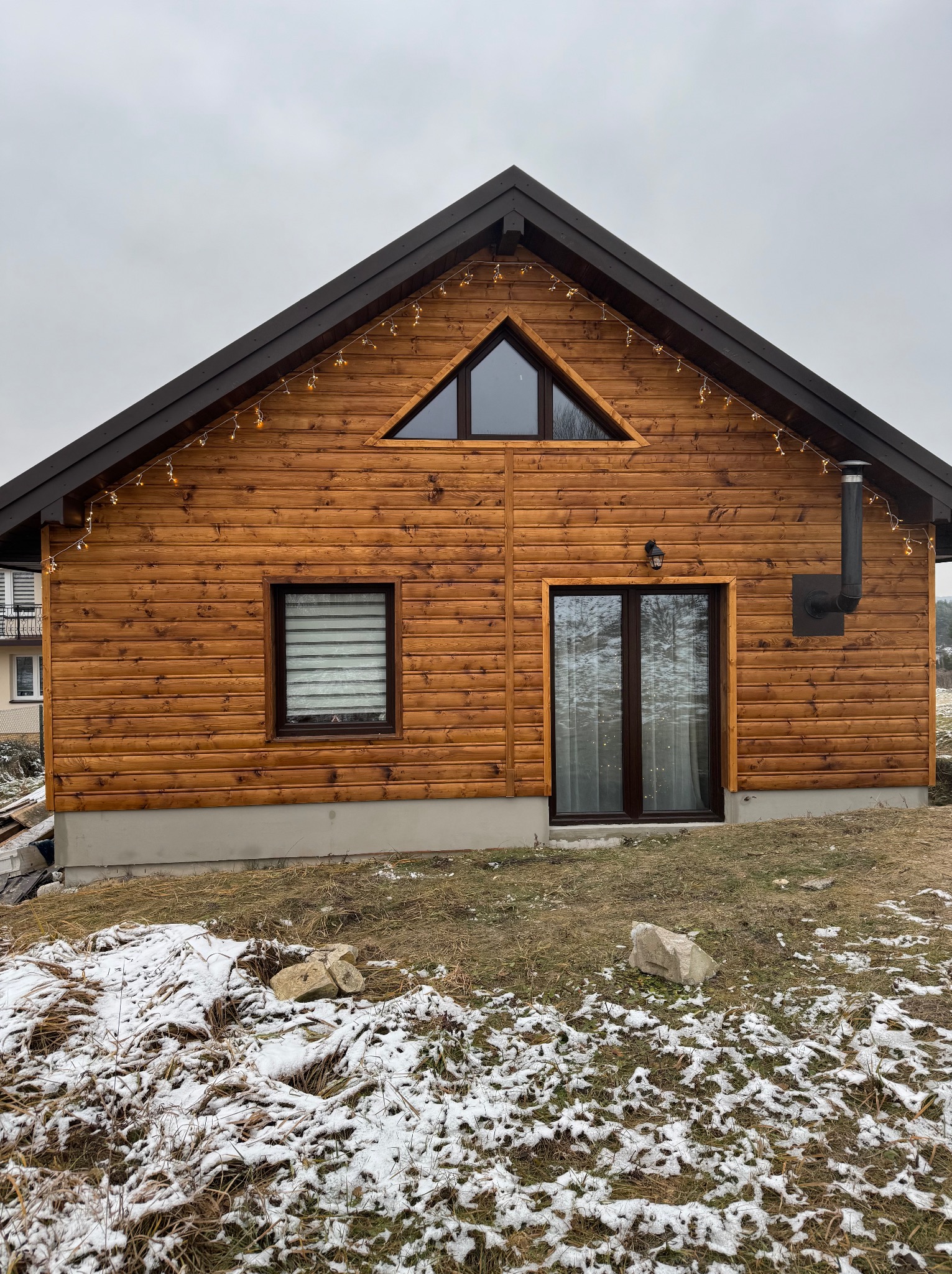 Drewniany dom z brązowym dachem i dekoracyjnymi lampkami, widoczny komin i okno z żaluzjami, częściowo ośnieżony teren.