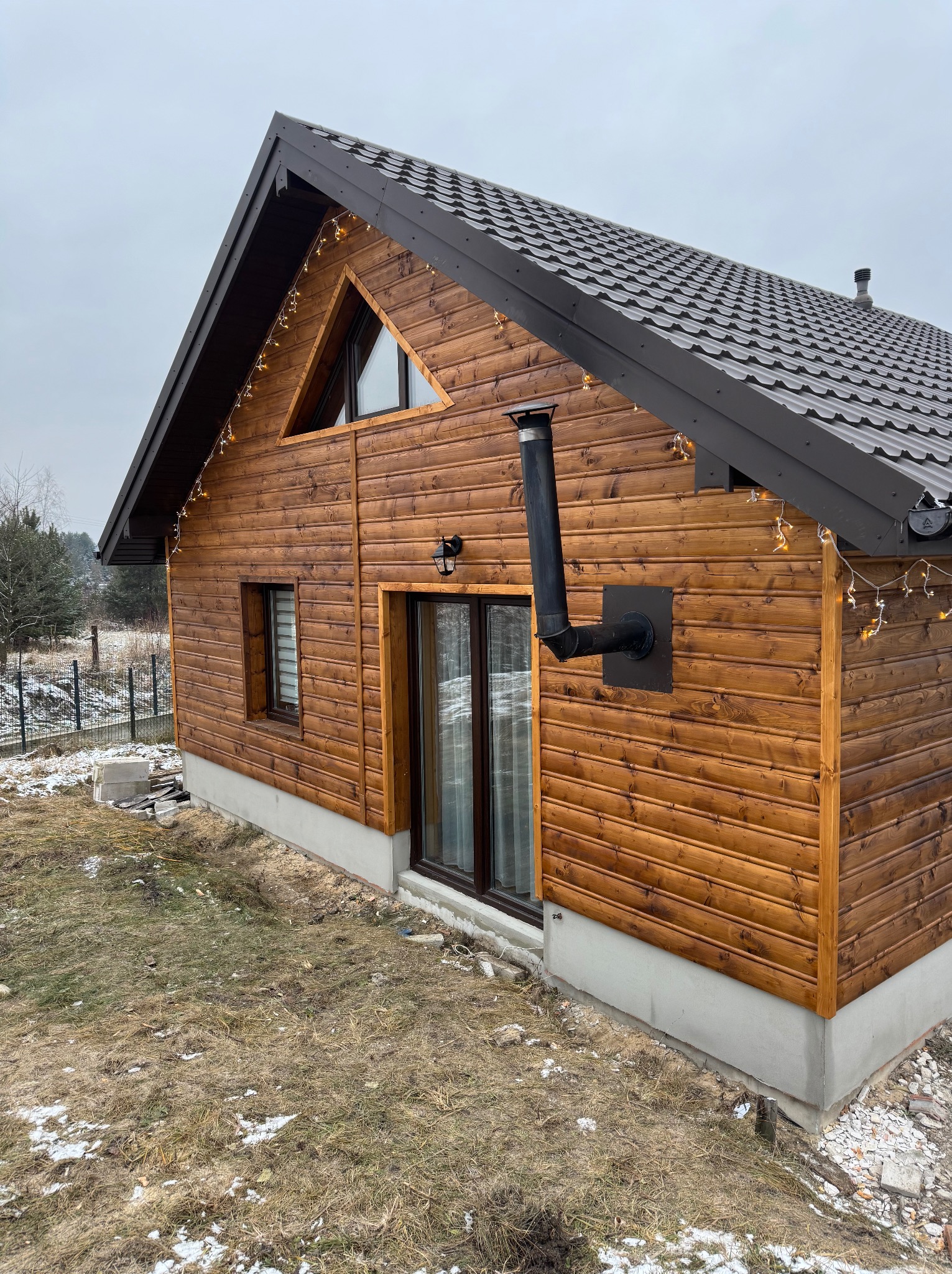 Drewniany dom z brązową blachodachówką, oświetleniem dekoracyjnym i czarnym kominem zewnętrznym, widok z zewnątrz na tle pochmurnego nieba.