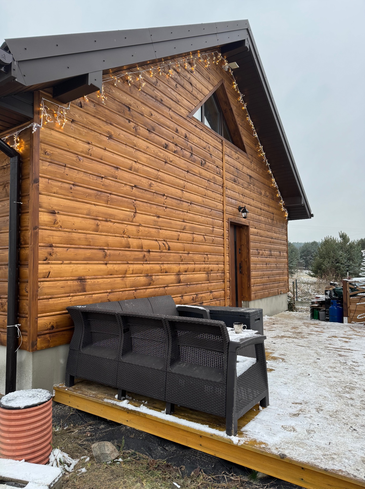 Drewniany dom z oknem w kształcie trójkąta, oświetlony girlandą lampek, z tarasem i meblami ogrodowymi w zimowej scenerii.