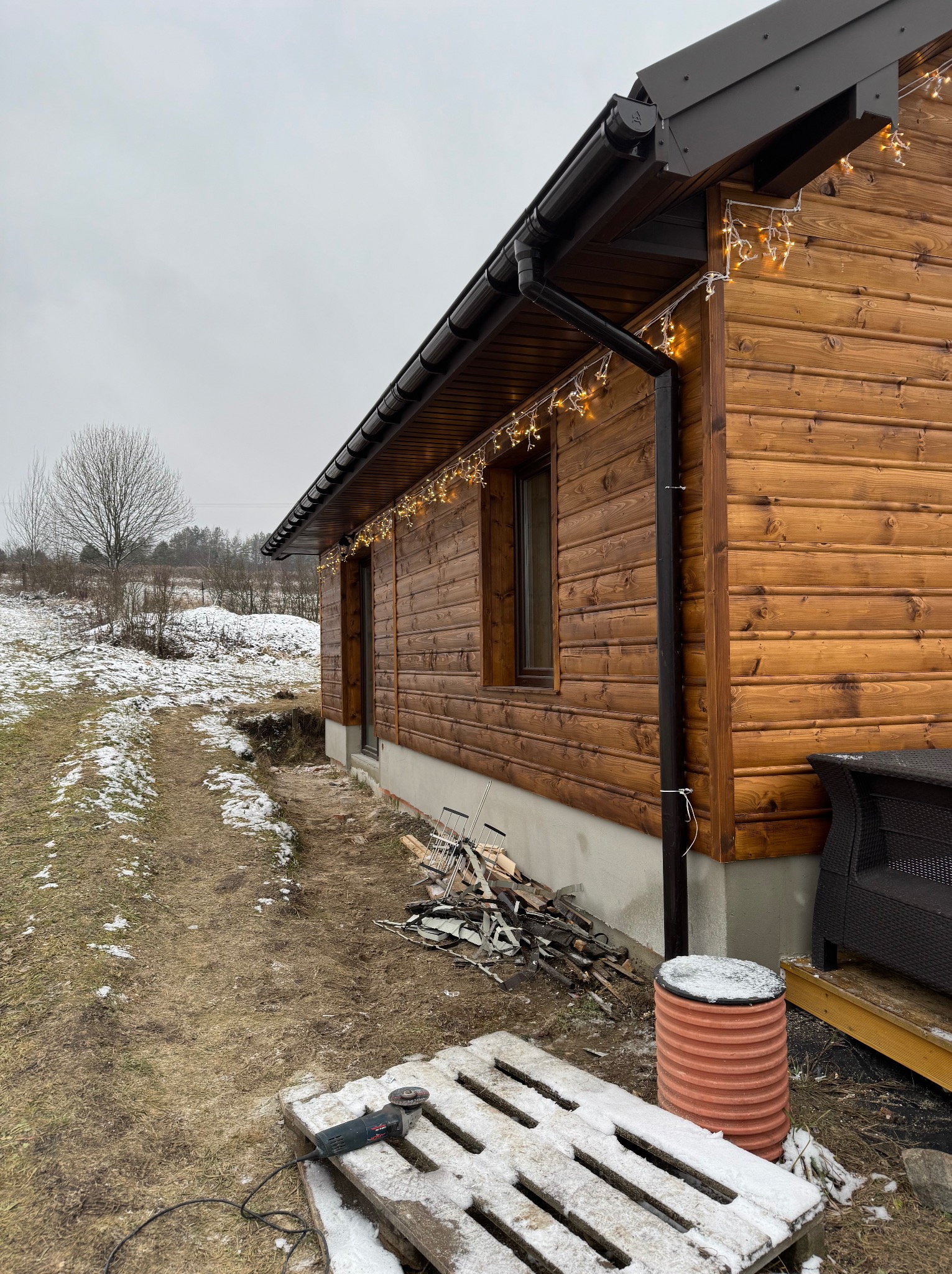 Ściana domu obita drewnem z zamontowanymi rynnami i dekoracyjnym oświetleniem, na ziemi paleta ze szlifierką kątową i resztki materiałów budowlanych.