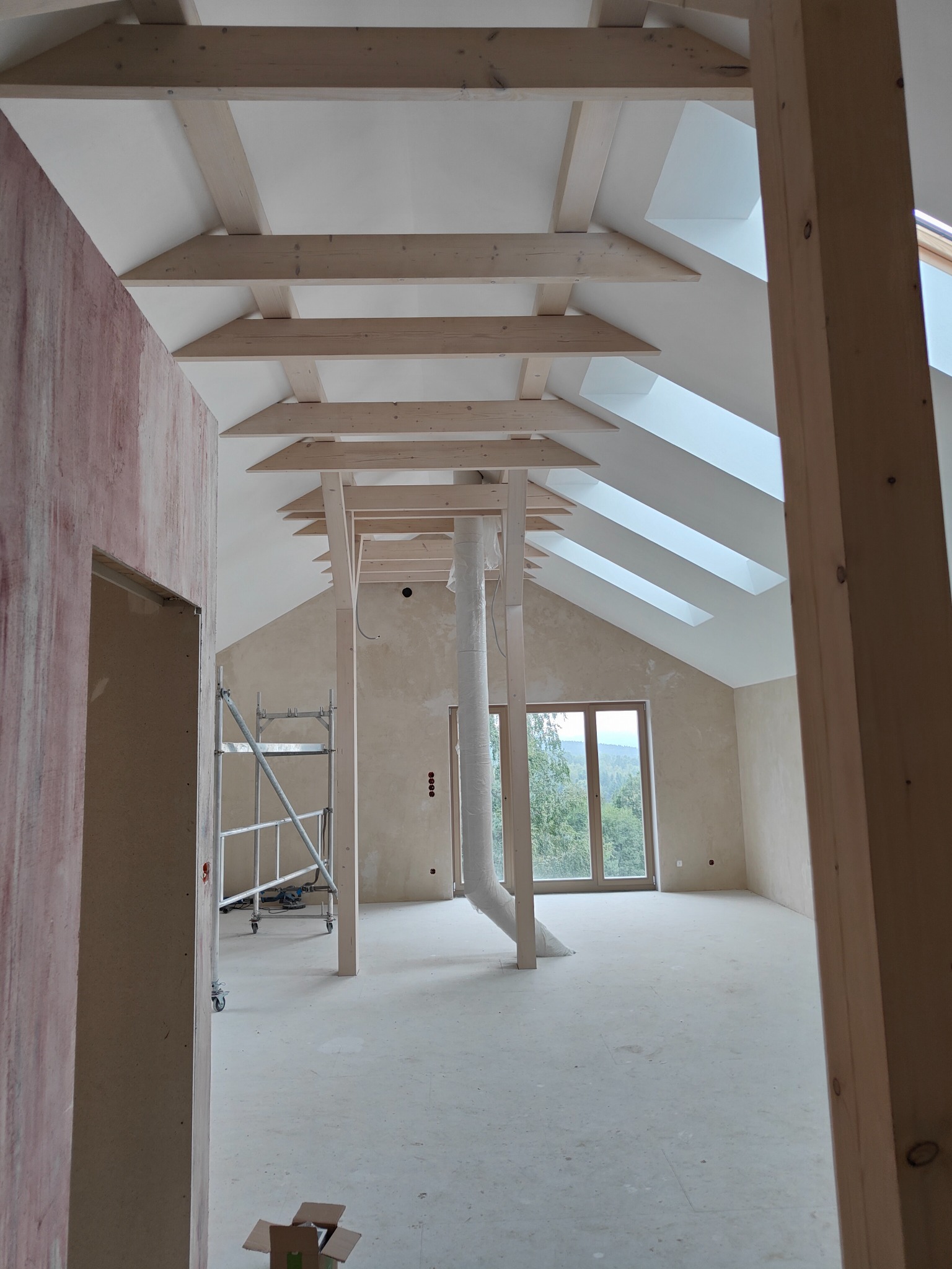 Poddasze w trakcie wykańczania, widoczne drewniane belki stropowe, okna dachowe, ściana z nałożonym tynkiem strukturalnym, rusztowanie i rura wentylacyjna.