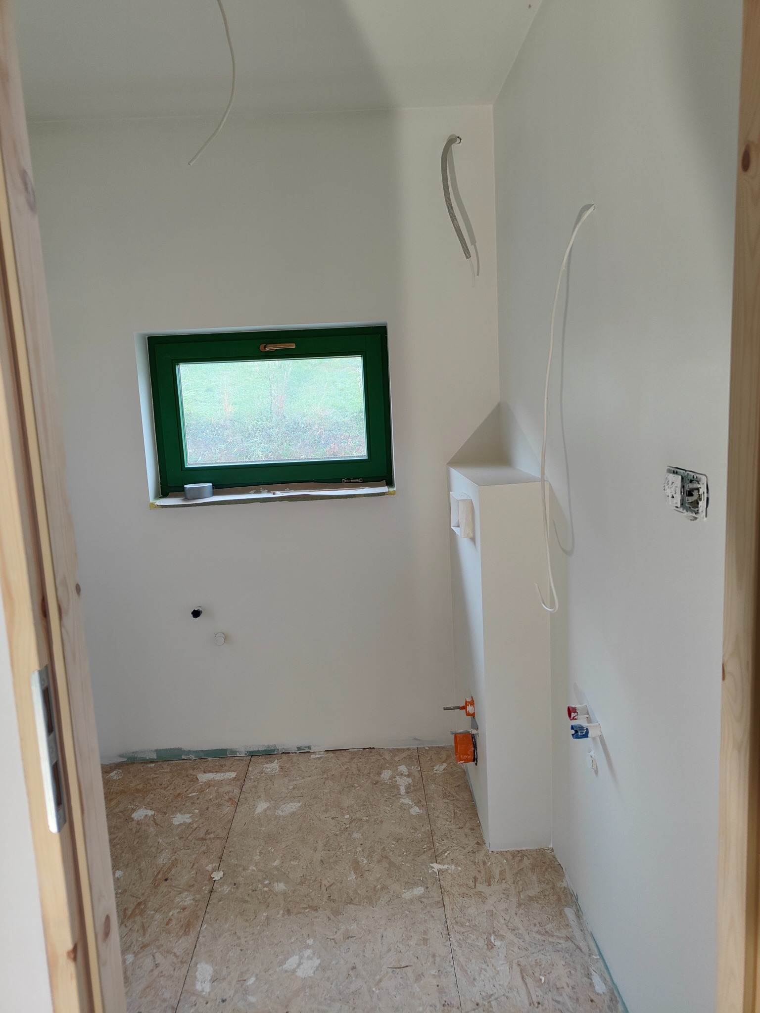 Wnętrze łazienki w trakcie remontu: surowe ściany pomalowane na biało, widoczne przewody elektryczne i instalacja wodna, podłoga z płyt OSB, zielone okno z widokiem na zewnątrz.