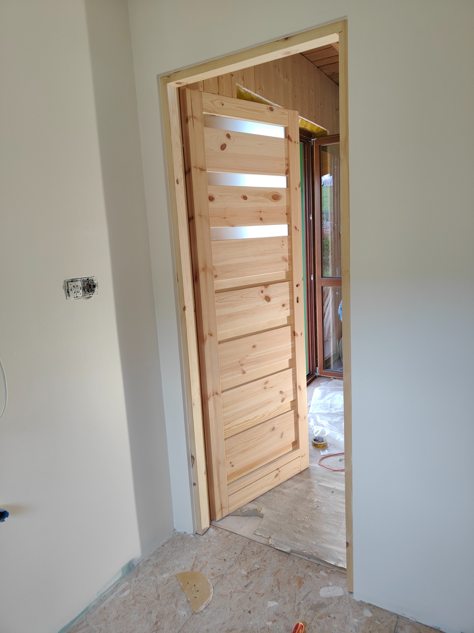 Wnętrze z częściowo zamontowanymi drewnianymi drzwiami z poziomymi przeszkleniami, widoczne niedokończone ściany i podłoga w trakcie remontu.