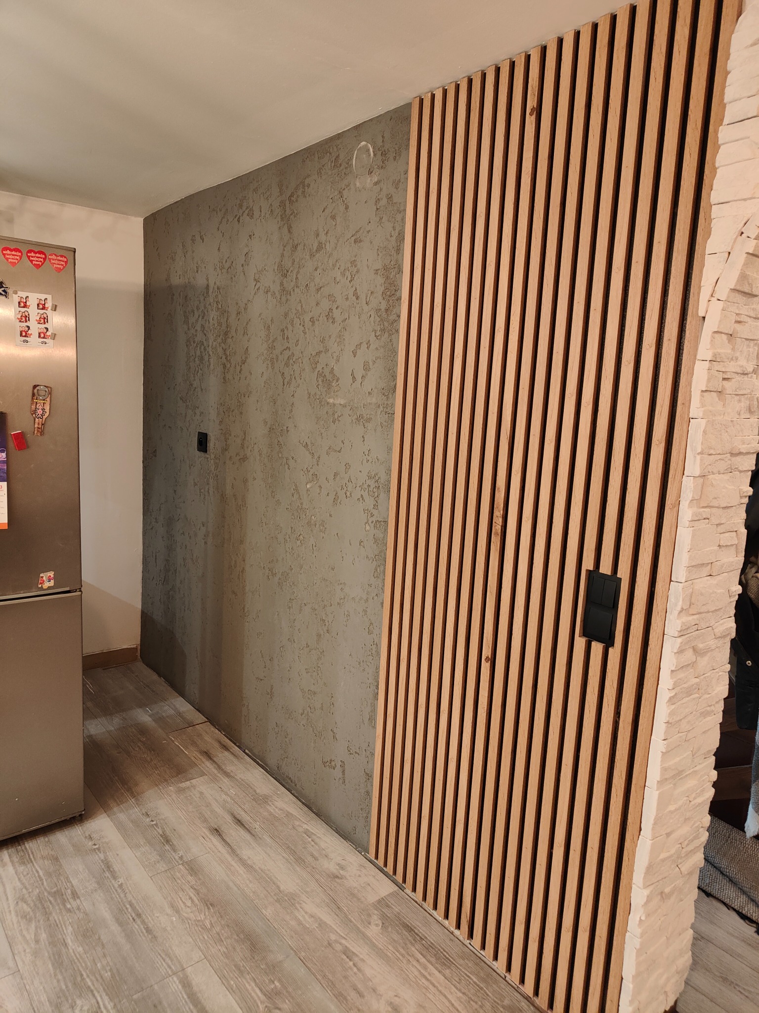 Dekoracyjna ściana z drewnianych lameli z włącznikiem światła, obok ściana z szarą fakturą i fragment lodówki ze zdjęciami na magnesach.