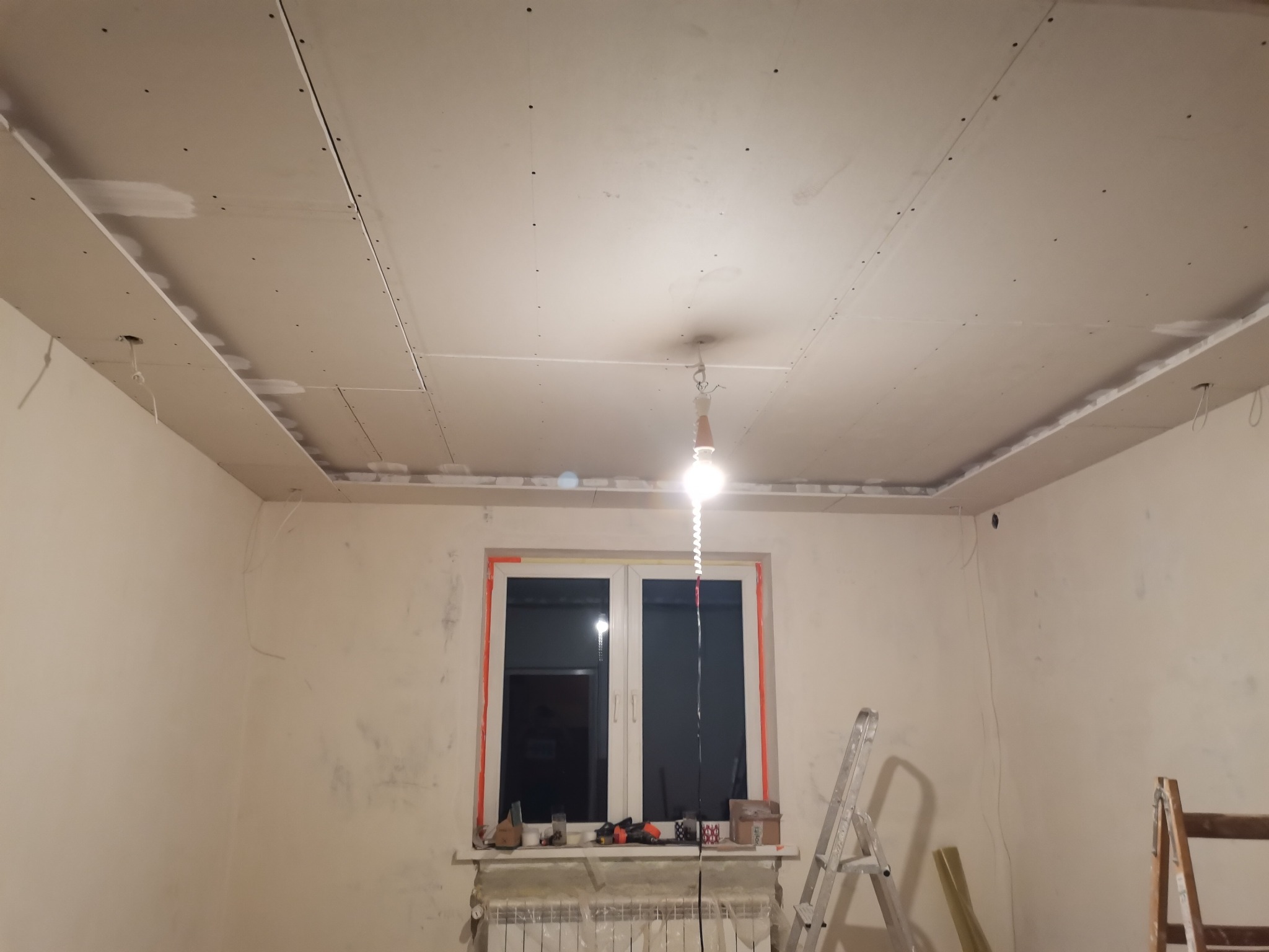 Montaż sufitu podwieszanego z płyty karton gips w trakcie realizacji, widoczne profile, śruby, szpachlowanie łączeń, prowizoryczne oświetlenie żarówką, drabiny i narzędzia na parapecie okiennym.