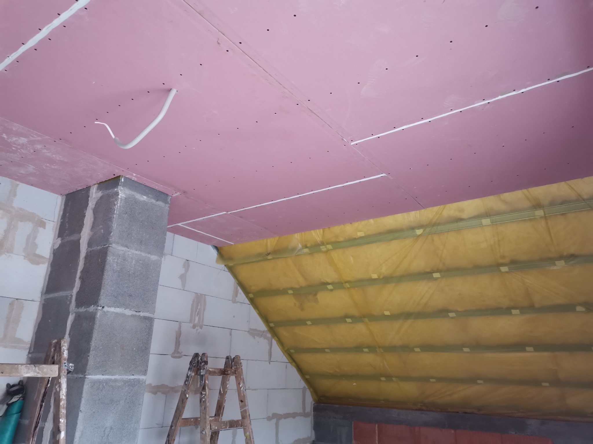 Poddasze w trakcie prac wykończeniowych: różowa płyta gipsowa na suficie, ocieplenie wełną mineralną na skosach, widoczne drabiny i fragment ściany z betonu komórkowego.