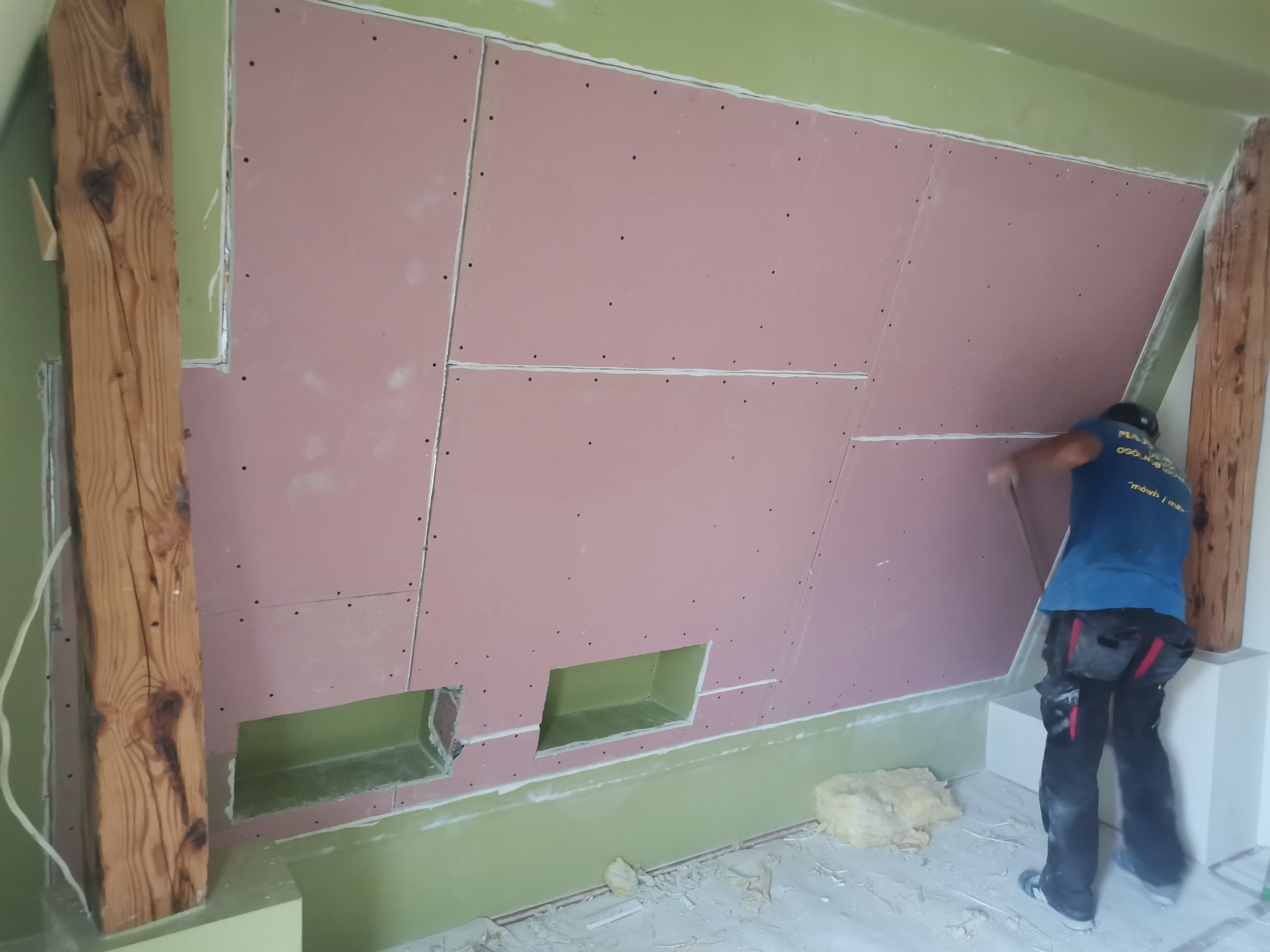 Montaż różowych płyt na poddaszu z widocznymi drewnianymi belkami konstrukcyjnymi i otworami na instalacje, wykonywany przez pracownika w niebieskiej koszulce.