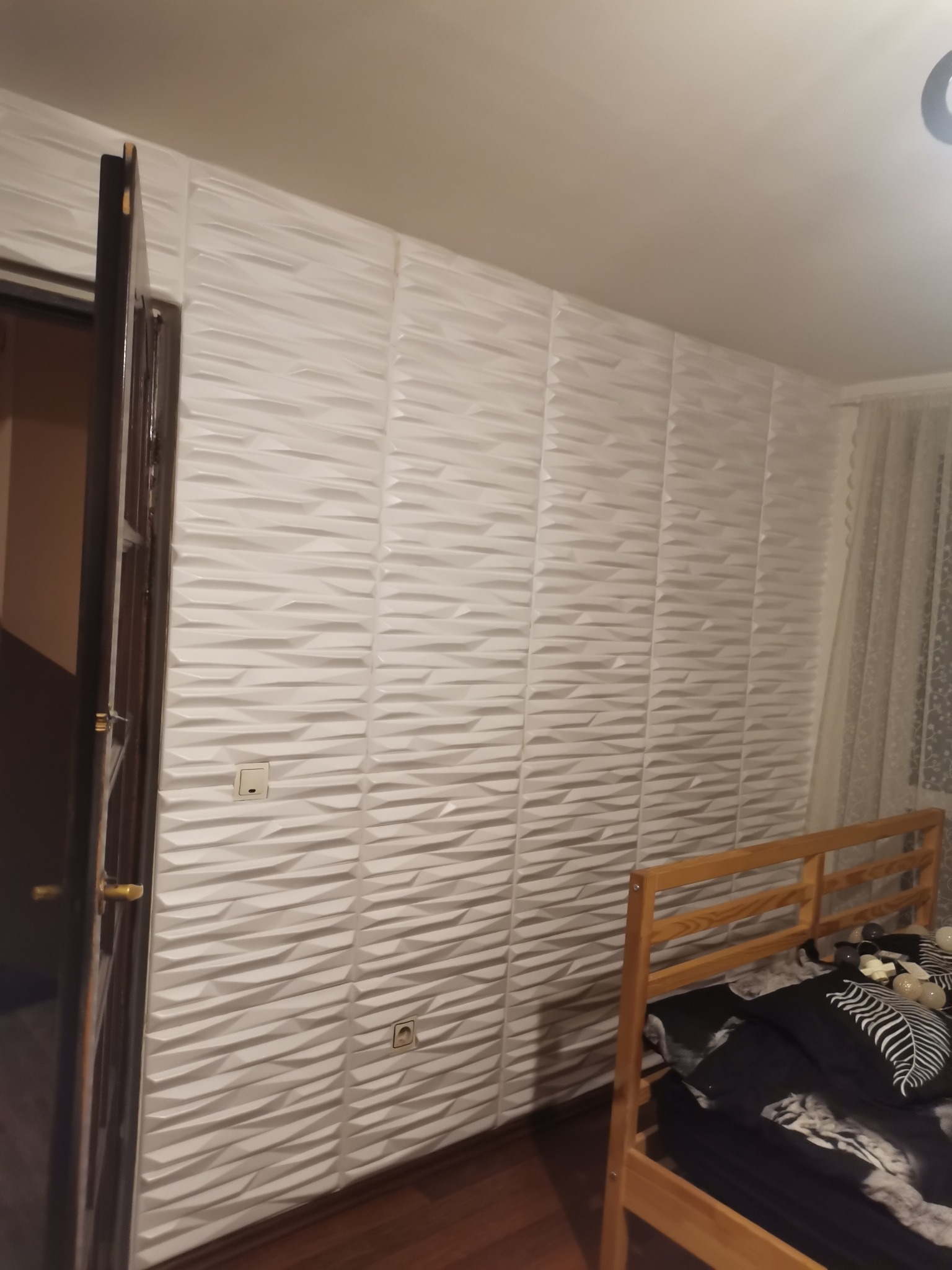 Ściana wykończona białymi panelami 3D z włącznikiem i gniazdkiem elektrycznym, widoczna drewniana rama łóżka i fragment otwartych drzwi.